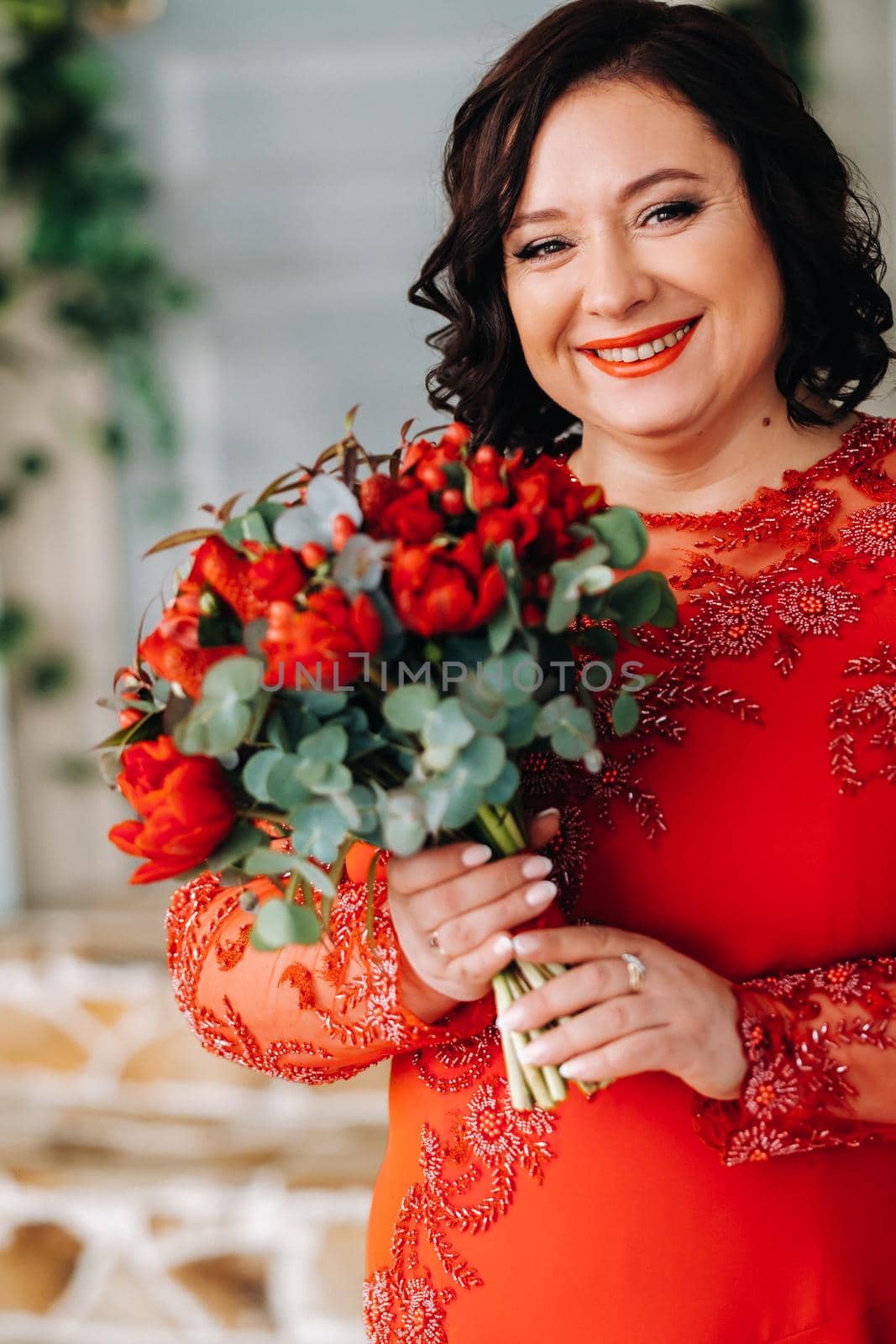 a woman in a red dress stands and holds a bouquet of red roses and strawberries in the interior