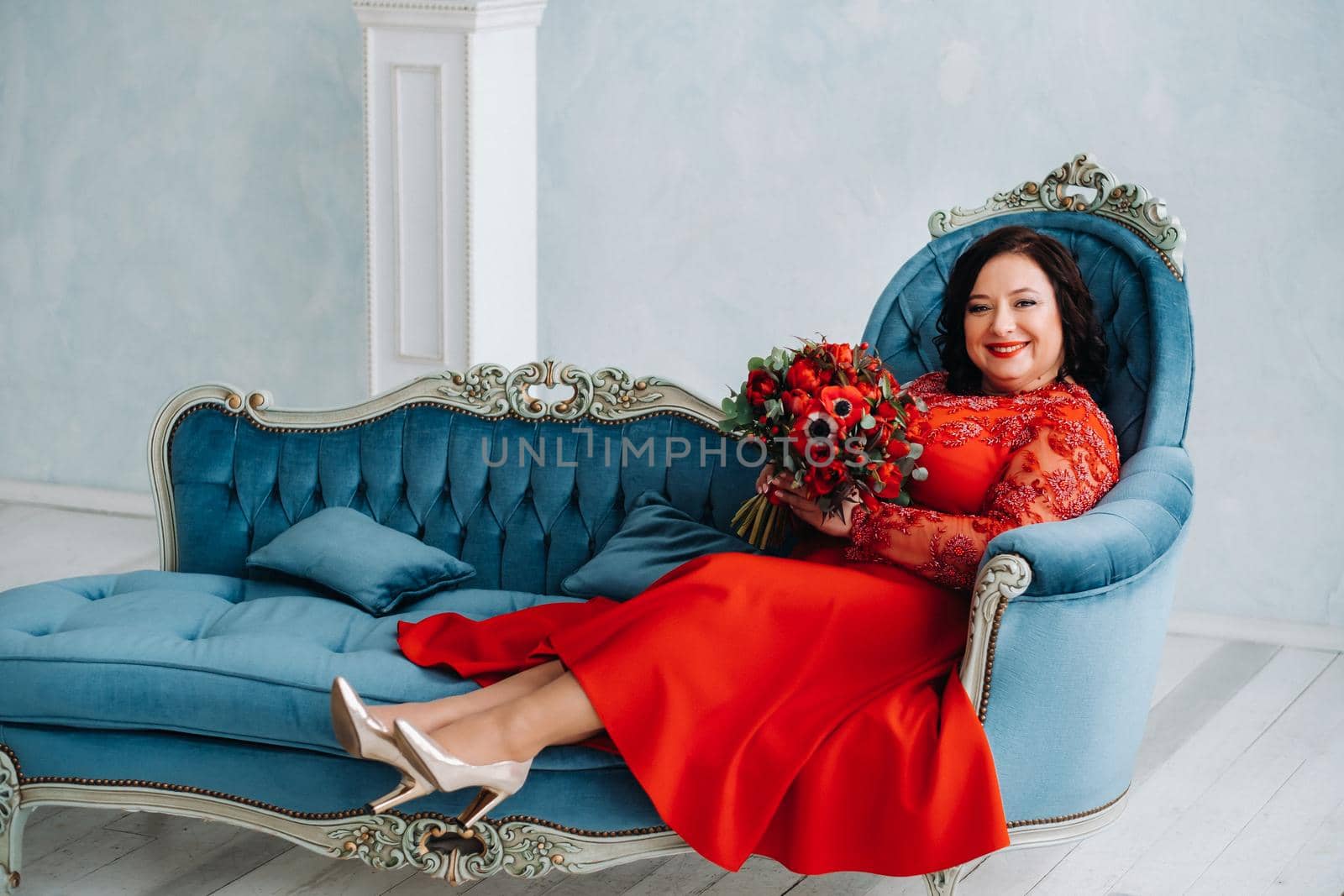 a woman in a red dress sits on a sofa and holds a bouquet of red roses and strawberries in the interior