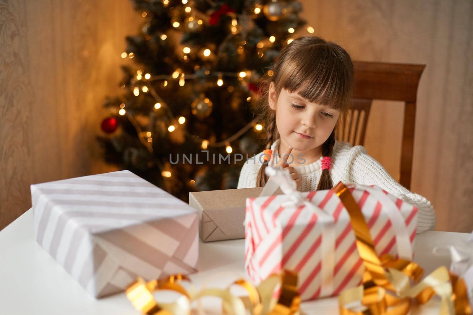 Charming child pack Christmas gifts in festive room, looks at boxes, wearing casual attire, having dark hair an two pigtails, presents for new year tree. by sementsovalesia