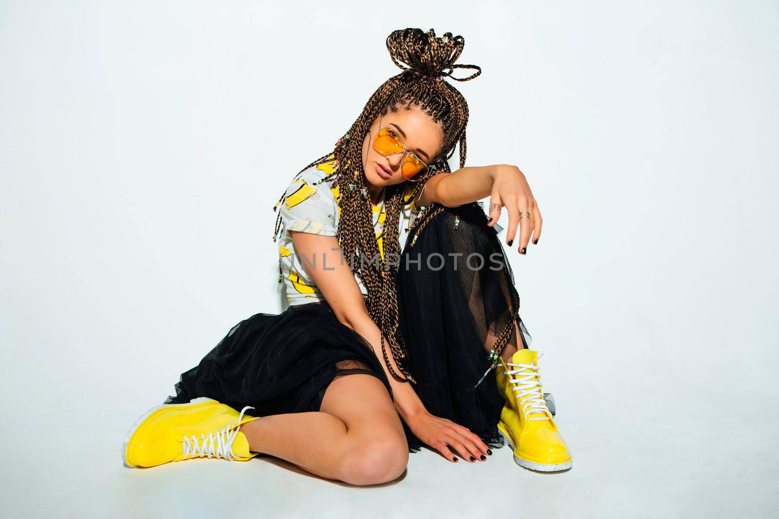 Portrait of a young stylish woman with braided hair dressed in white t-shirt, black skirt, yellow boots and sunglasses posing on the white background.