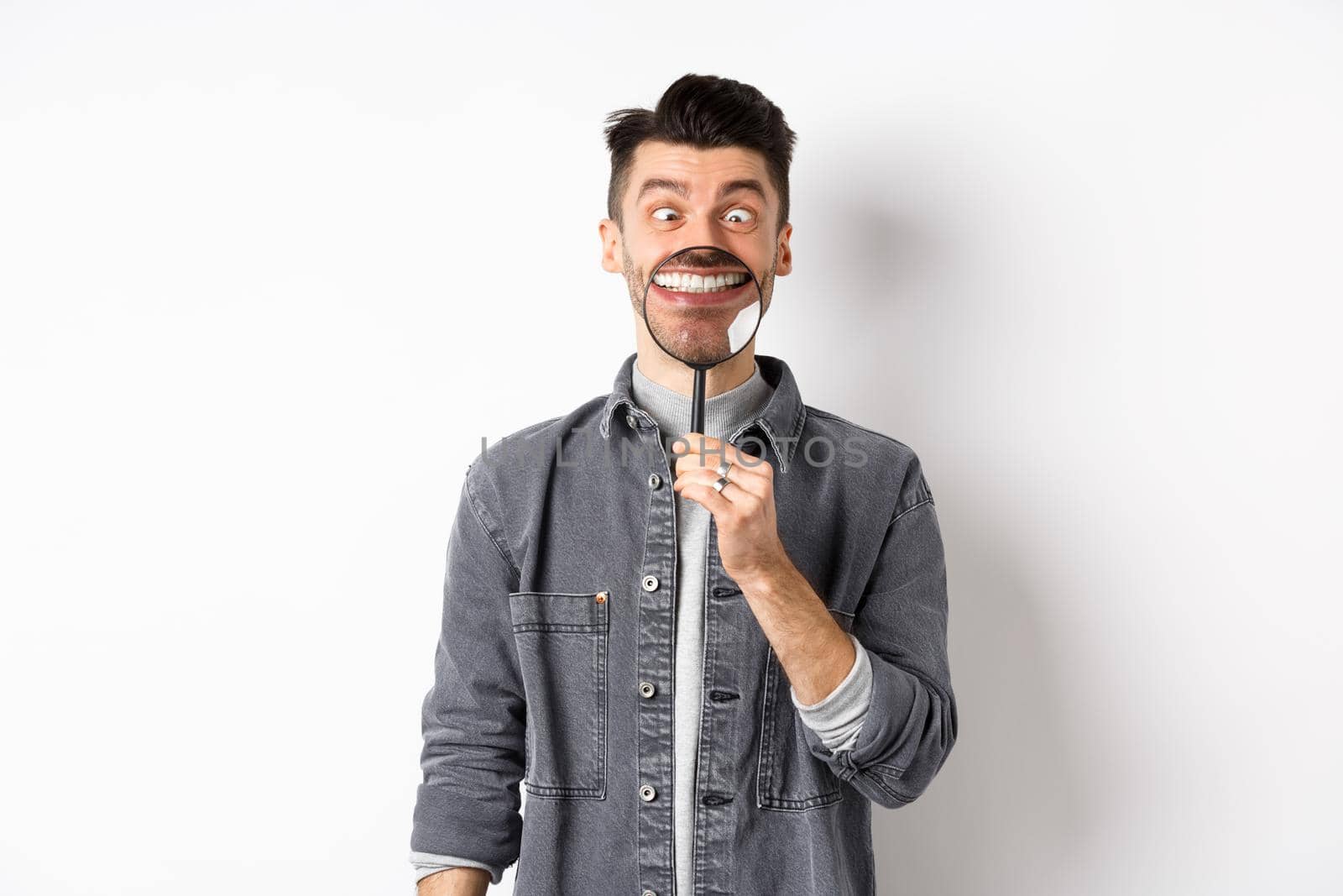 Handsome positive guy showing white perfect smile with magnifying glass, and squinting eyes, making funny faces, white background.