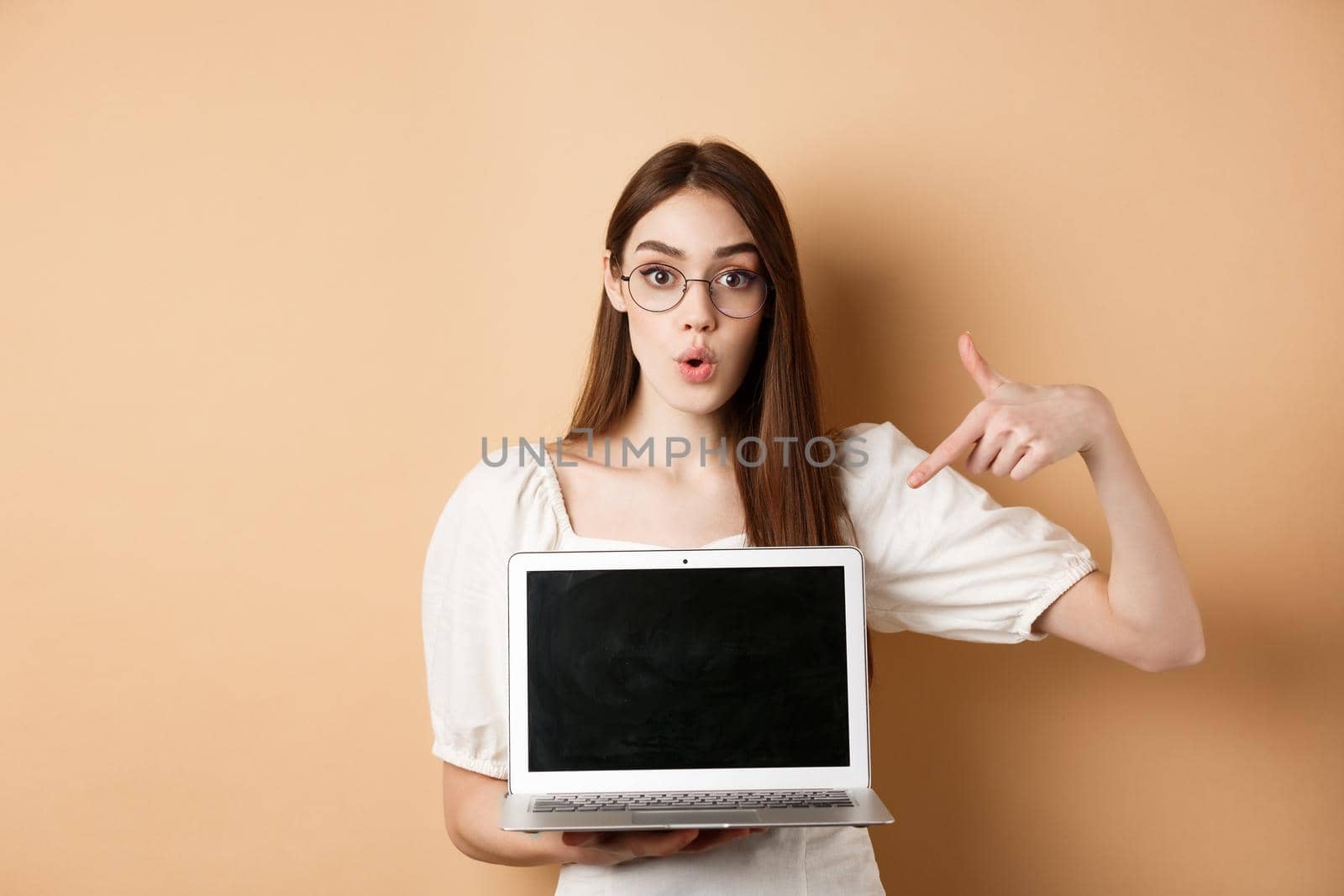 Excited girl in glasses making presentation on computer, pointing hand at laptop screen and say wow, standing on beige background by Benzoix