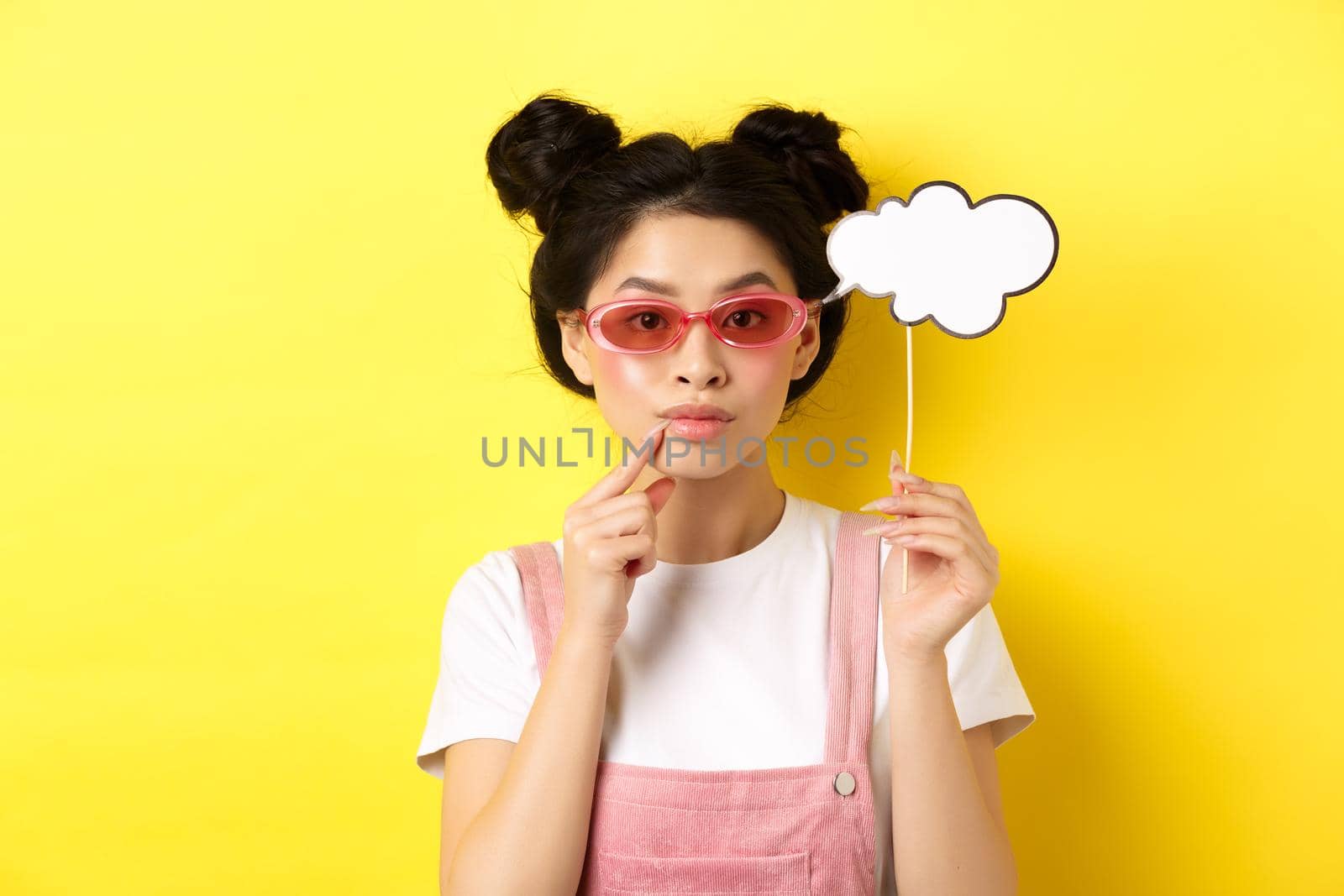 Summer and fashion concept. Stylish asian girl in sunglasses holding comment cloud on stick and looking thoughtful, thinking or saying something, standing on yellow background.