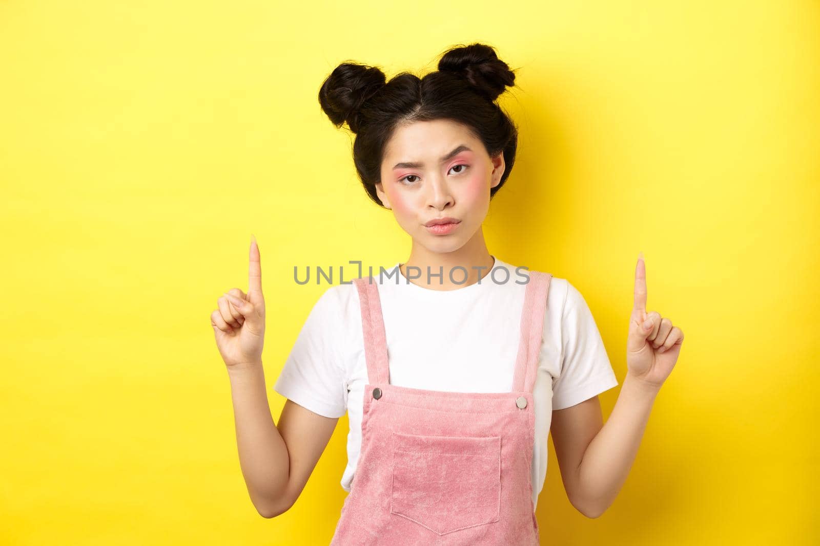 Annoyed and tired asian girl with pink glamour makeup, pointing fingers up and look bothered, standing on yellow background by Benzoix