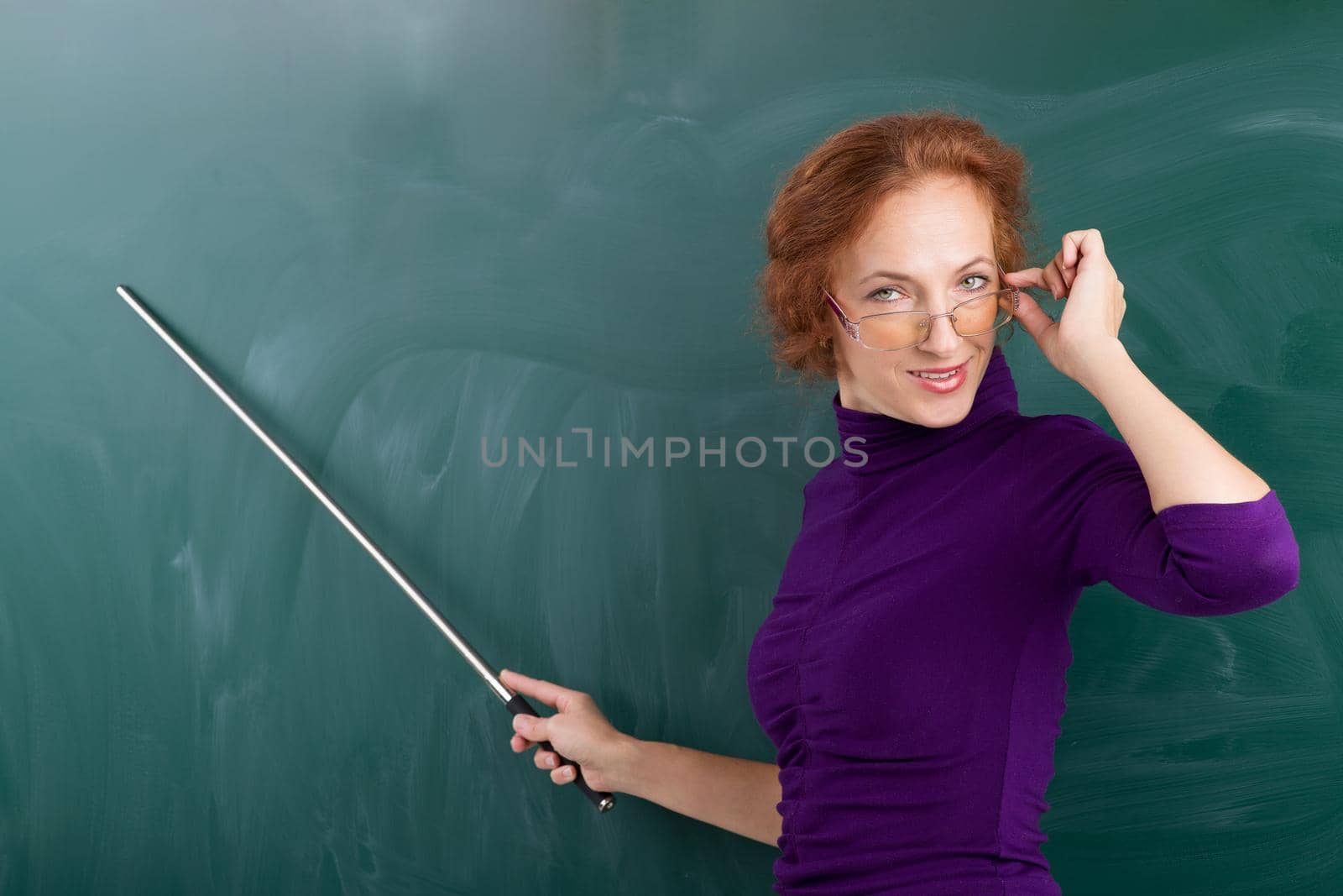 Teacher standing at blackboard with pointer. Portrait of smiling young woman looking over glasses. Teacher posing at green chalkboard and pointing at it