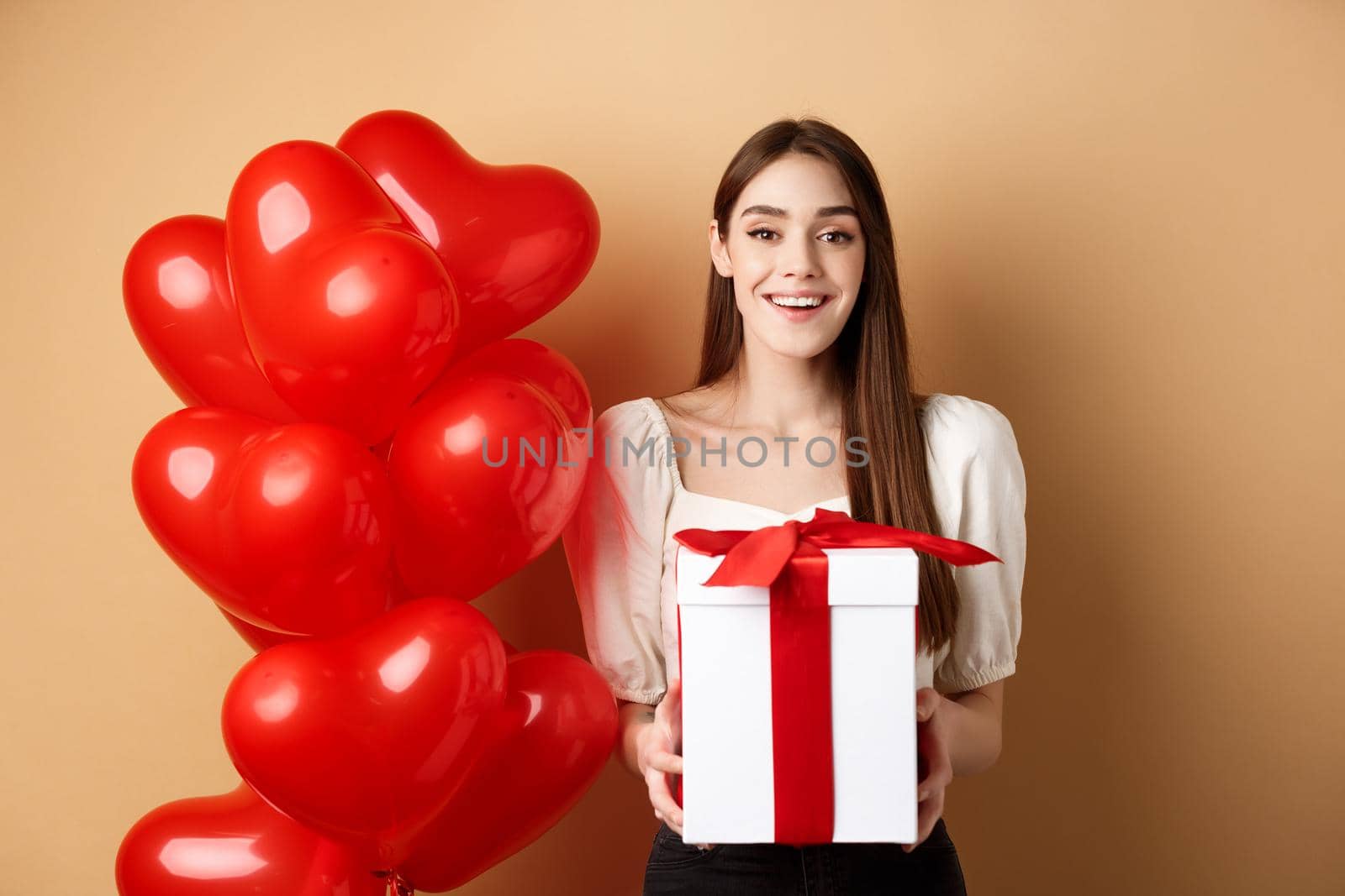 Romantic girl standing near heart balloons and holding surprise gift on Valentines day, smiling at camera happy, standing on beige background by Benzoix