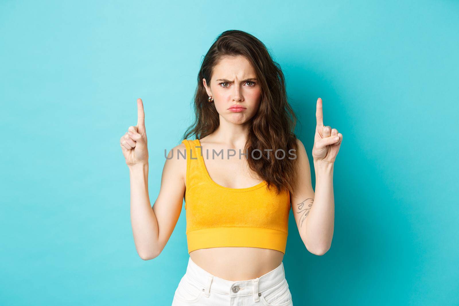 Summer lifestyle concept. Disappointed moody girl complaining about unfair situation, frowning and sulking upset, pointing fingers up at logo, standing over blue background.