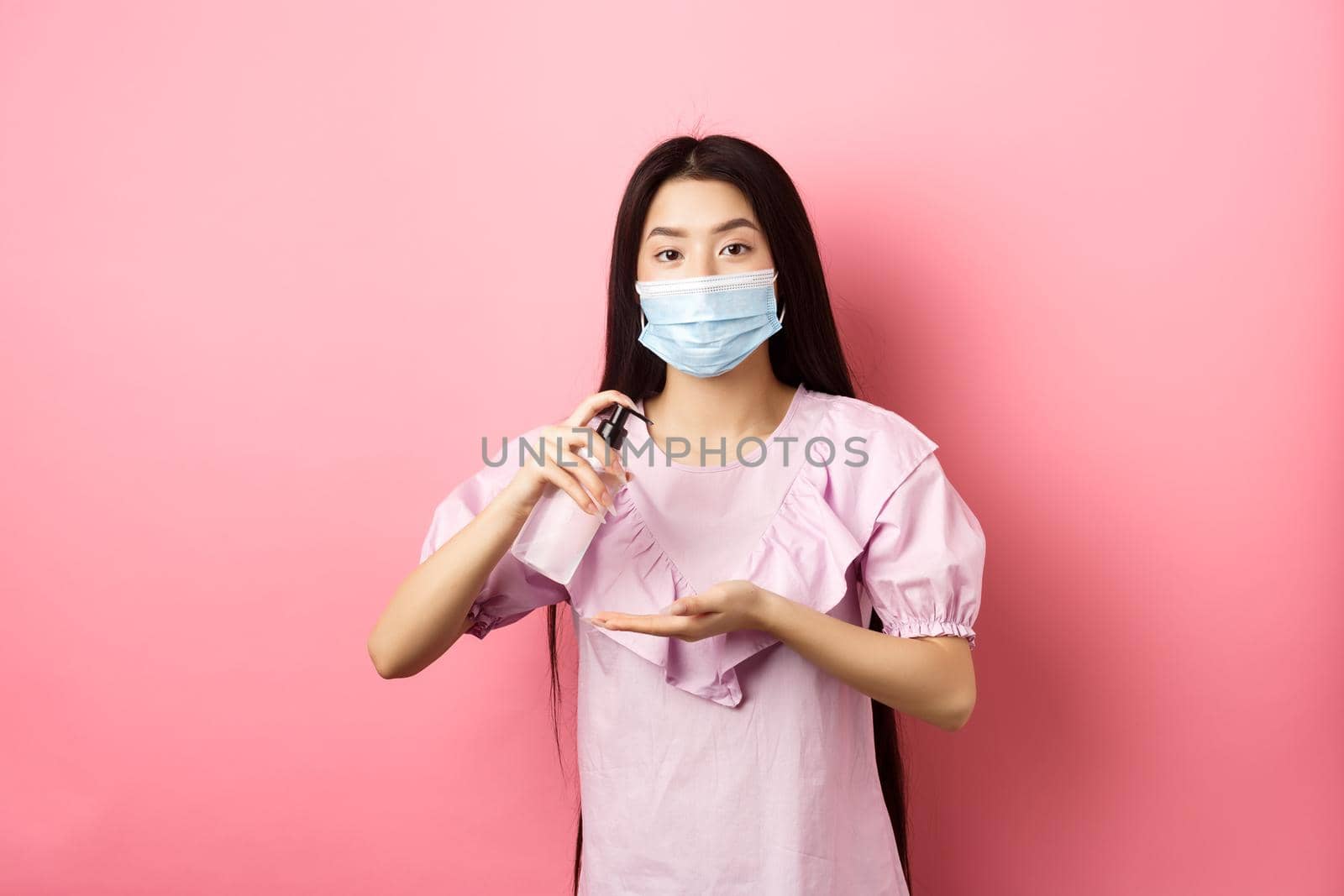 Healthy people and covid-19 pandemic concept. Cute asian woman in medical mask clean hands with antiseptic, using sanitizer, standing against pink background by Benzoix