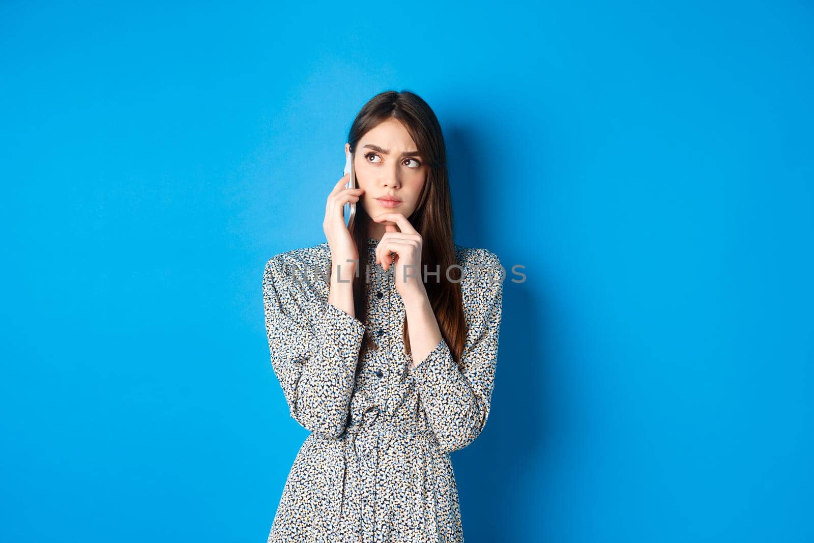Pensive woman talking on phone and thinking, looking aside thoughtful, standing against blue background by Benzoix