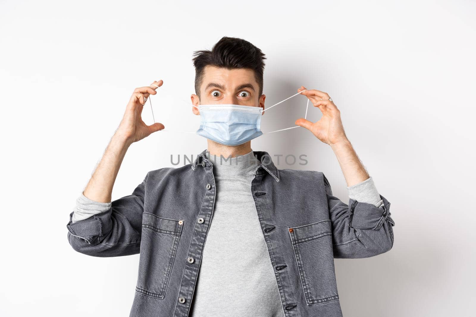 Health, covid and pandemic concept. Funny guy put on medical mask and look at camera, prevent catching coronavirus, standing on white background.