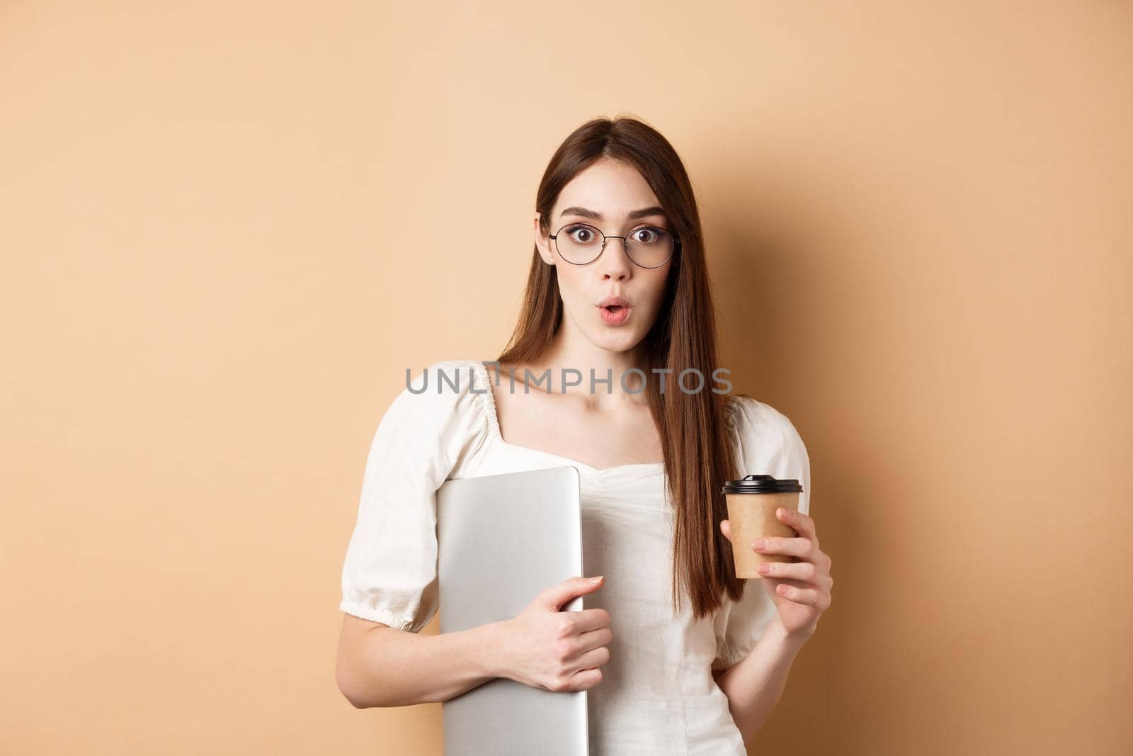 Working woman in glasses holding laptop and coffee, looking excited, hear amazing news, standing on beige background.