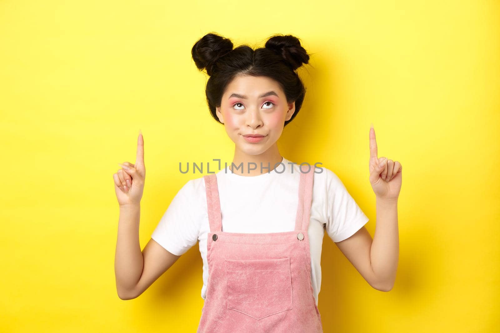 Glamour asian girl with beauty makeup, pointing fingers up, showing top advertisement, standing on yellow background.