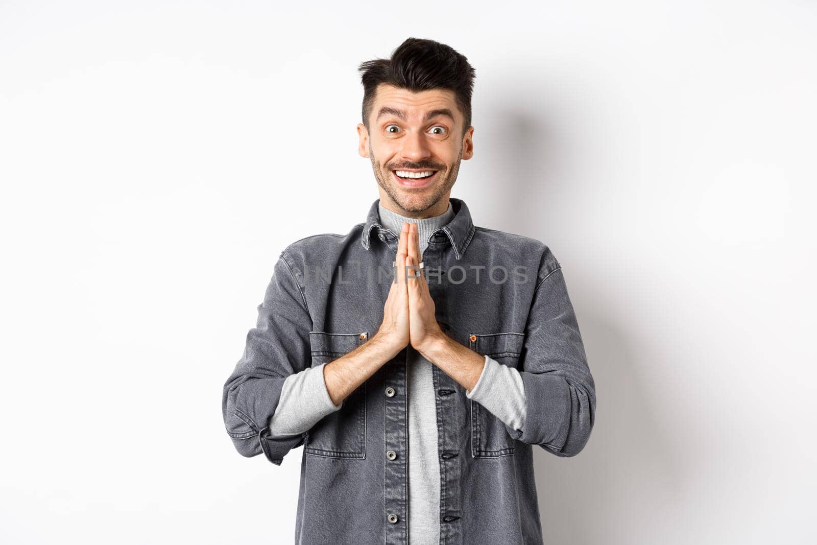 Please help me. Smiling cute man holding hands in namaste gesture, asking for favour, saying thank you, looking hopeful and excited at camera, standing on white background.