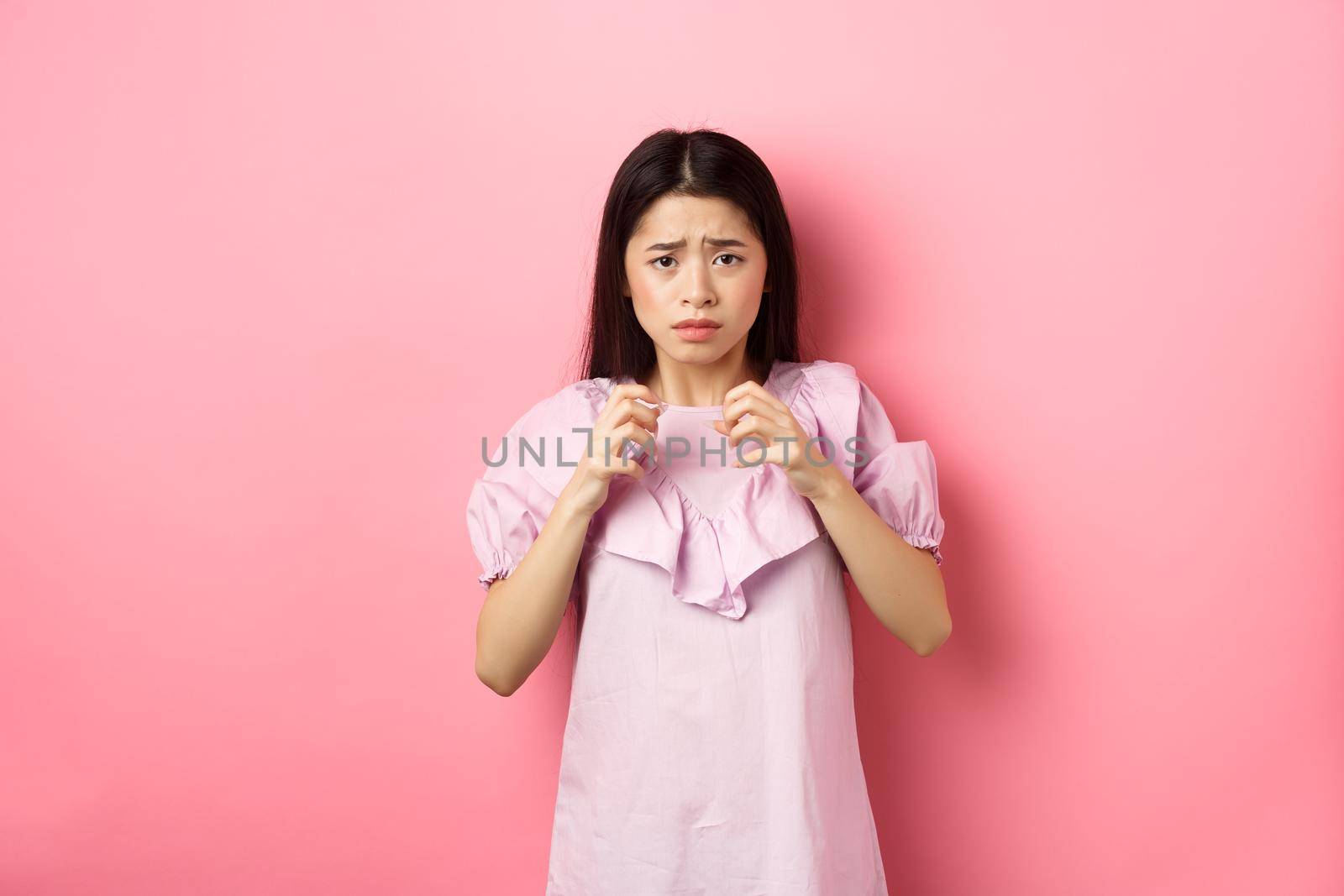 Scared asian woman victim, looking frightened, trembling from fear, stare at scary thing, standing anxious on pink background by Benzoix