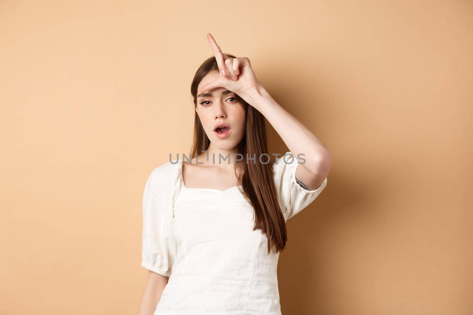 Confident woman mocking people with loser sign, being mean, standing on beige background.