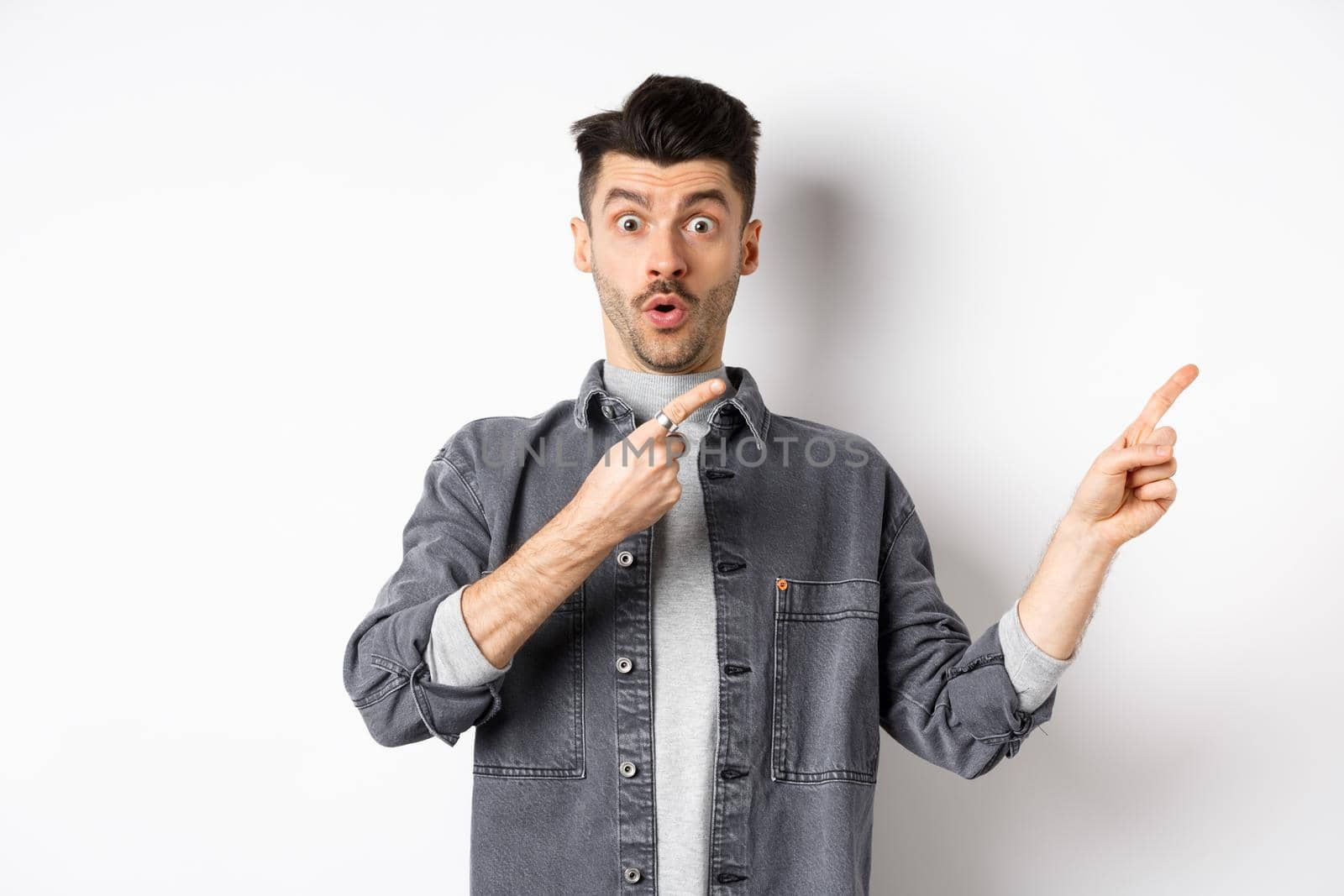 Excited guy showing advertisement aside, pointing fingers right and gasping amazed, checking out special deal, standing on white background by Benzoix