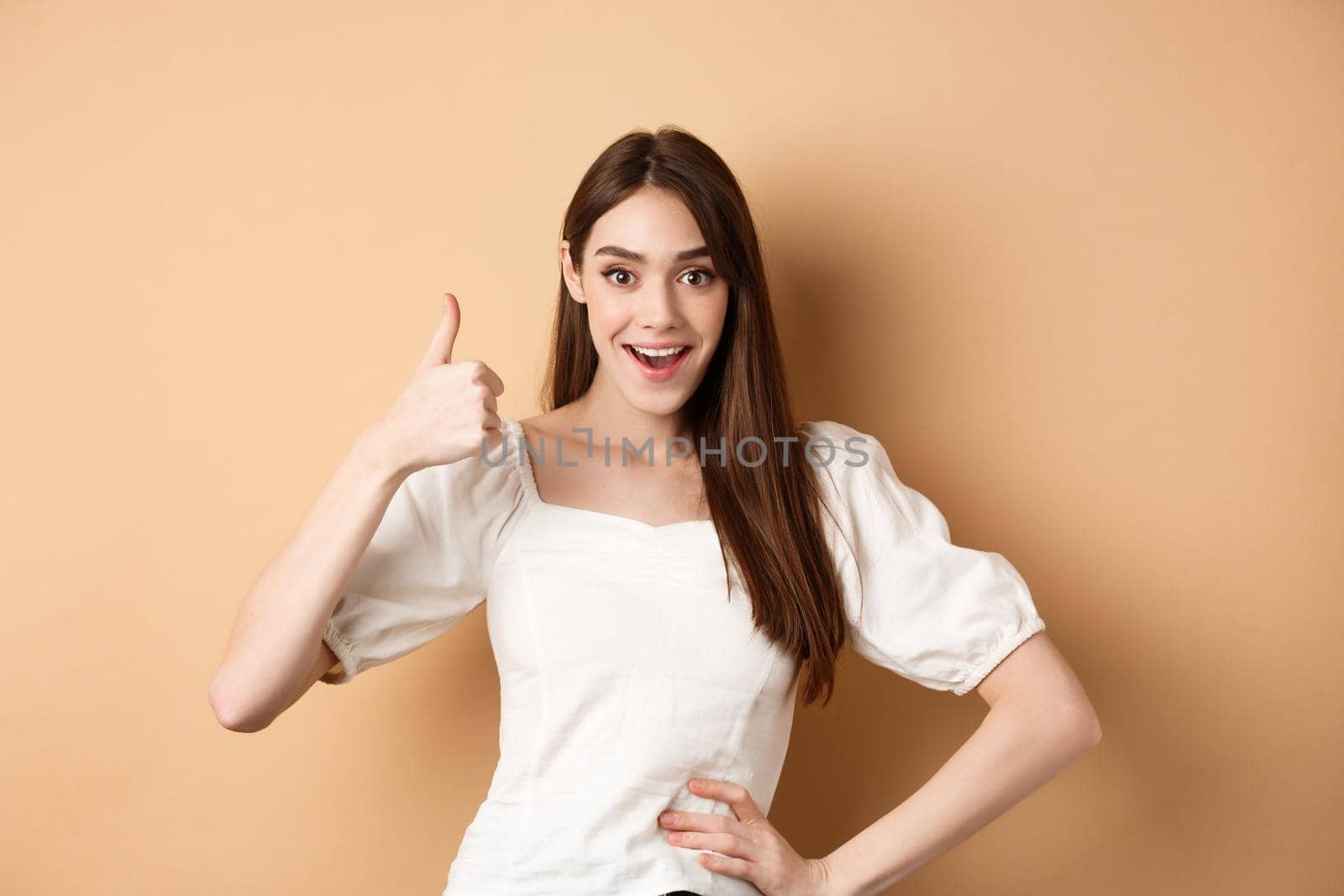 Amazed young woman showing thumb up, say yes and praise great product, approve choice, standing satisfied on beige background.
