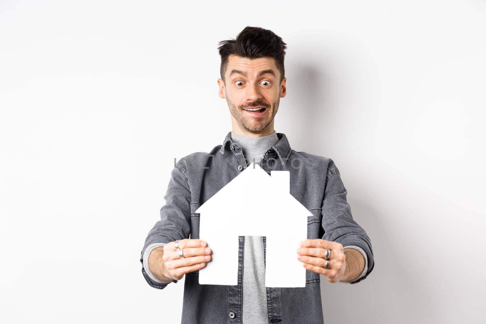 Real estate and insurance concept. Man look with excitement at paper house cutout, buying home, standing on white background.