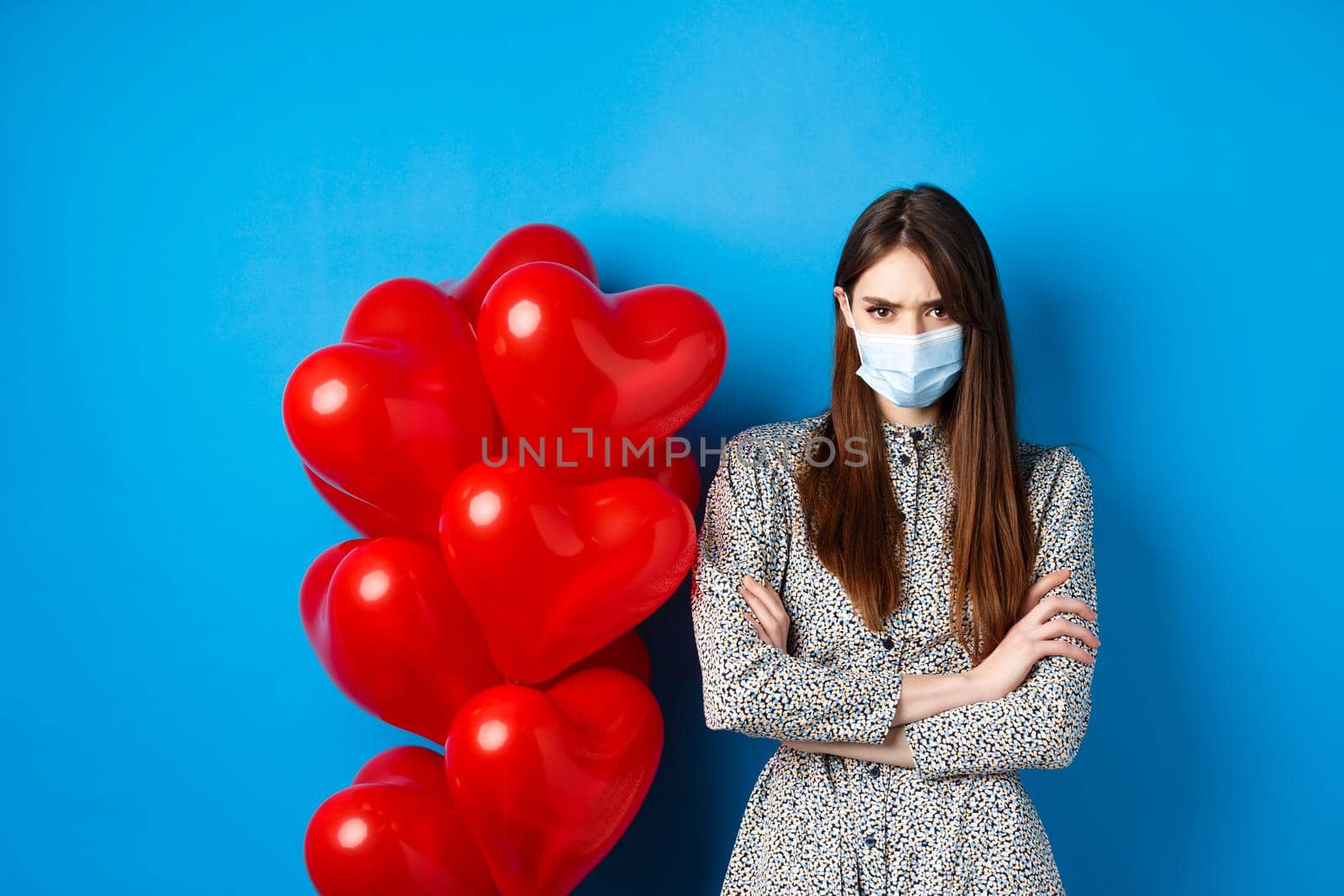 Covid-19, quarantine and health conept. Angry girlfriend in medical mask, standing near Valentines day red heart balloons, cross arms on chest and frowning offended, blue background by Benzoix