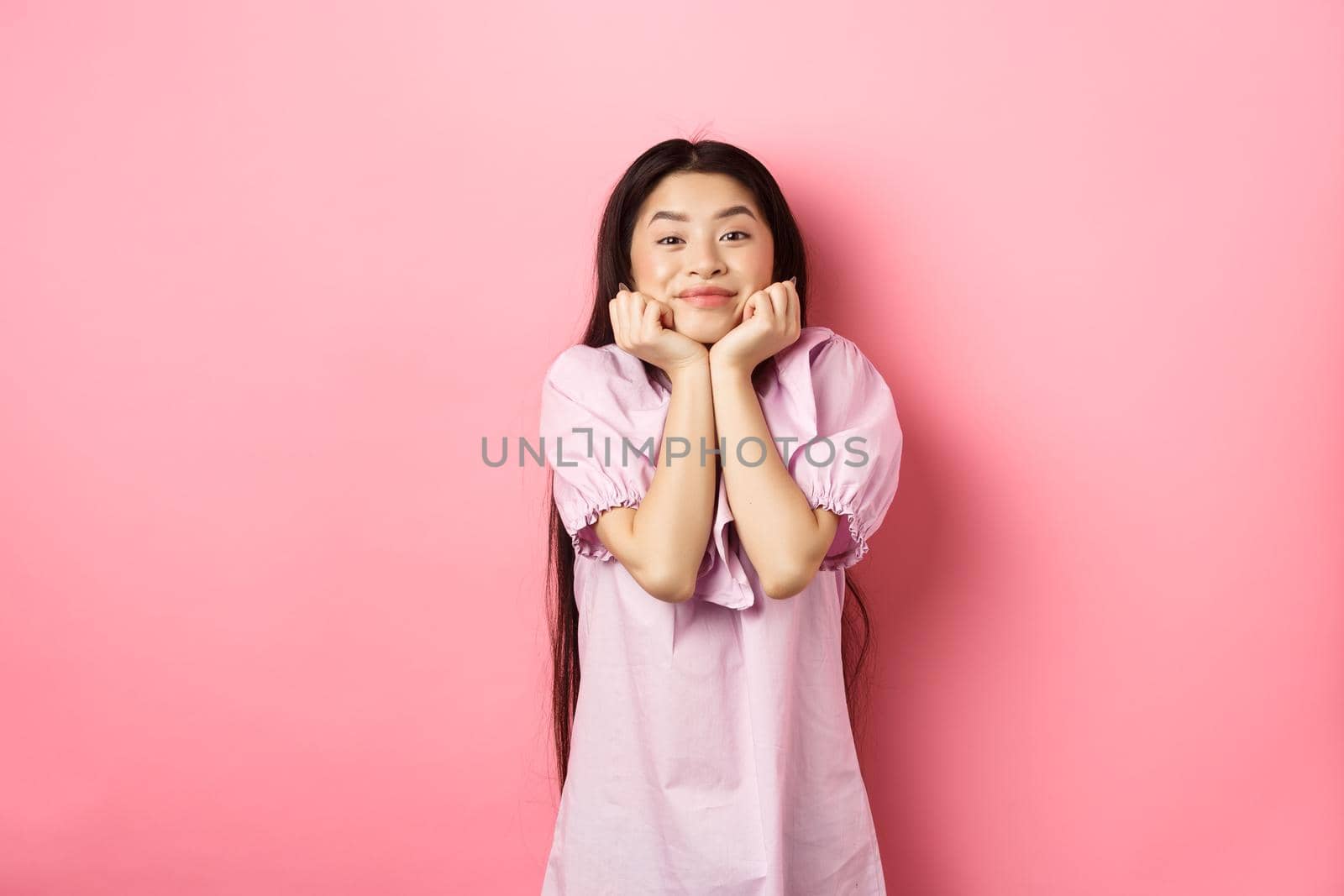 Cute and happy asian woman waiting for something with excitement, lean face on hands and watching lovely scene with admiration, standing against pink background by Benzoix