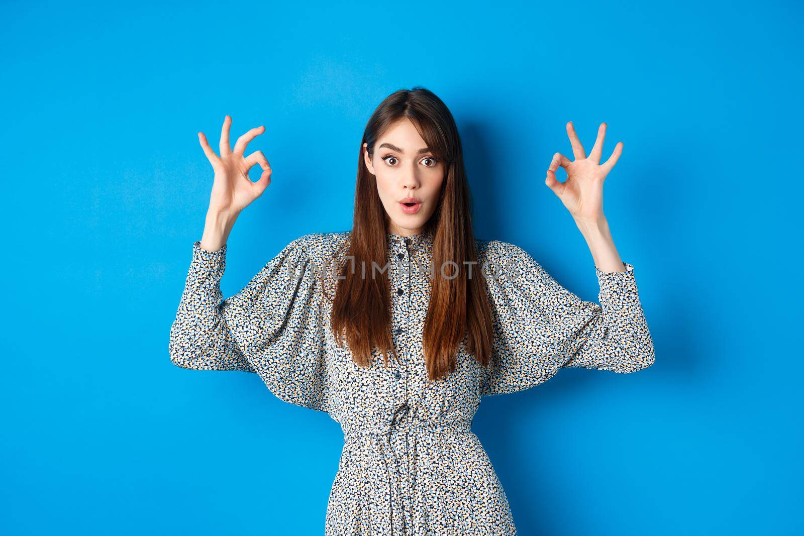 Excited beautiful woman in dress say wow, recommend cool thing, praising and looking impressed, standing on blue background by Benzoix