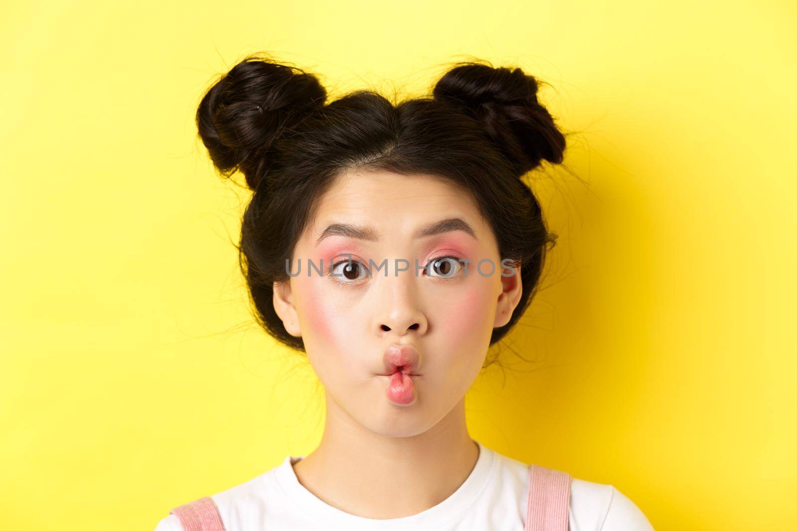 Close-up of teen korean girl pucker lips and looking funny at camera, standing with glamour makeup and stylish hairstyle, yellow background.