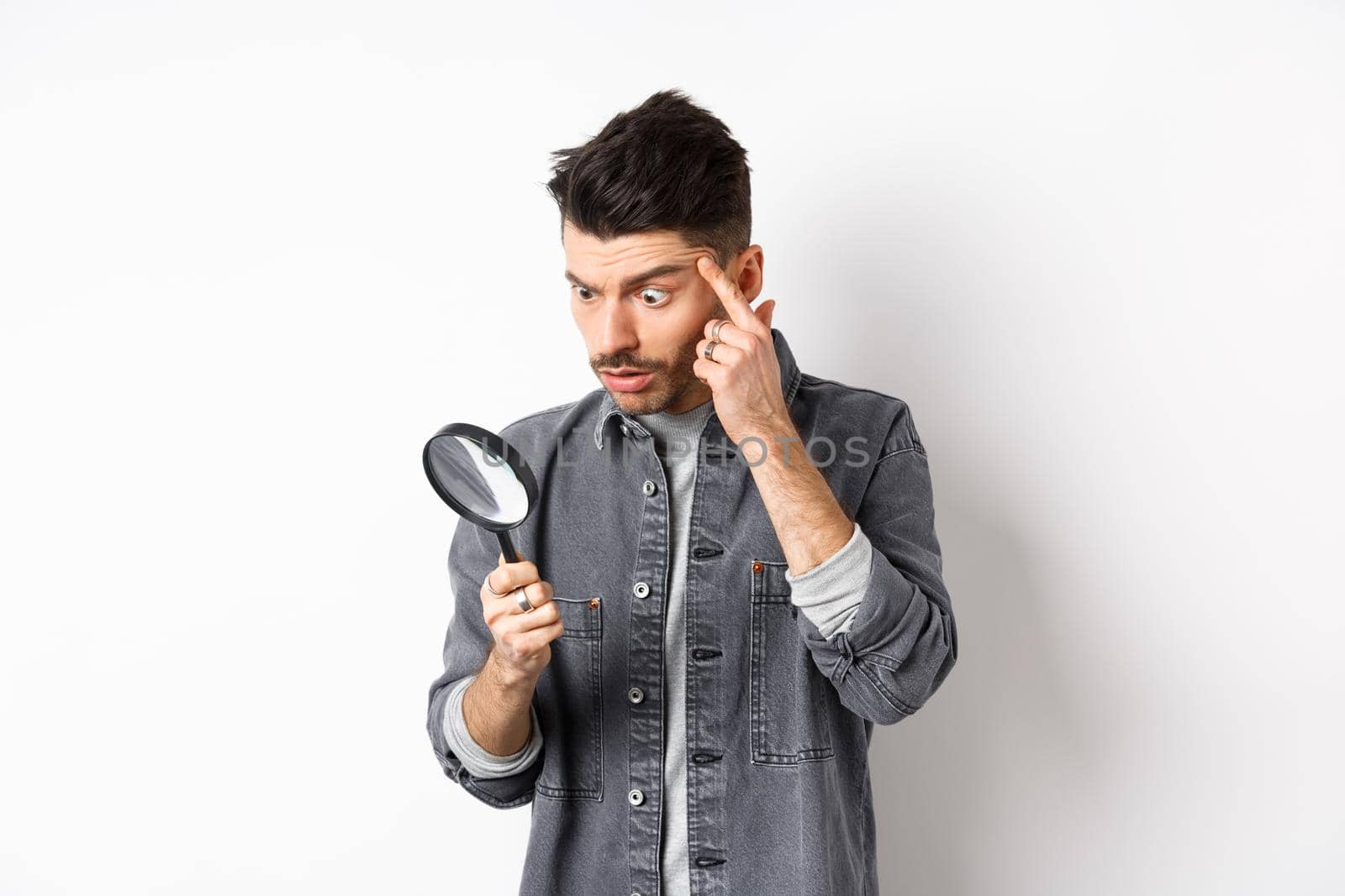 Man looking through magnifying glass and stretching eyelid to see clearly, standing on white background.