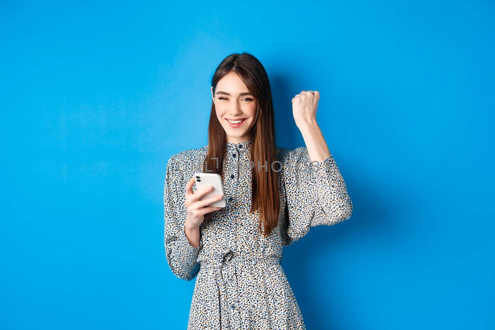 Happy pretty girl winning on mobile phone, smiling and chanting, holding smartphone, standing in dress against blue background by Benzoix