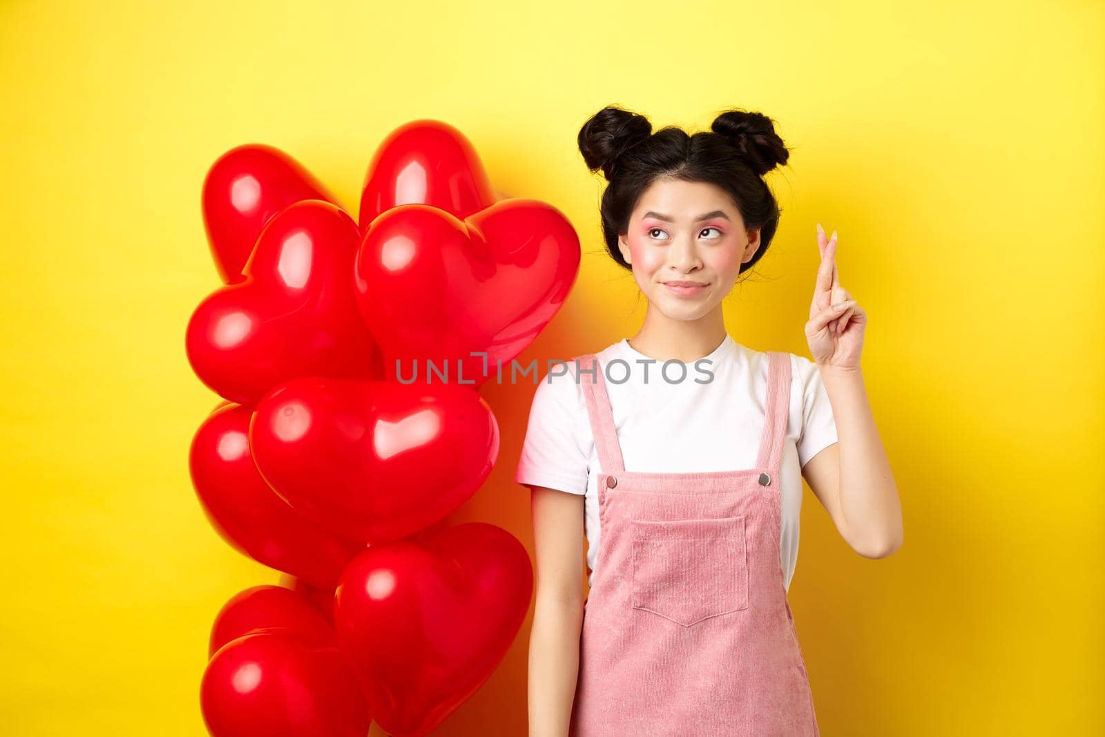 Valentines day concept. Beautiful korean girl dreaming of perfect date, making wish and looking at upper left corner, standing near red hearts balloons, yellow background by Benzoix