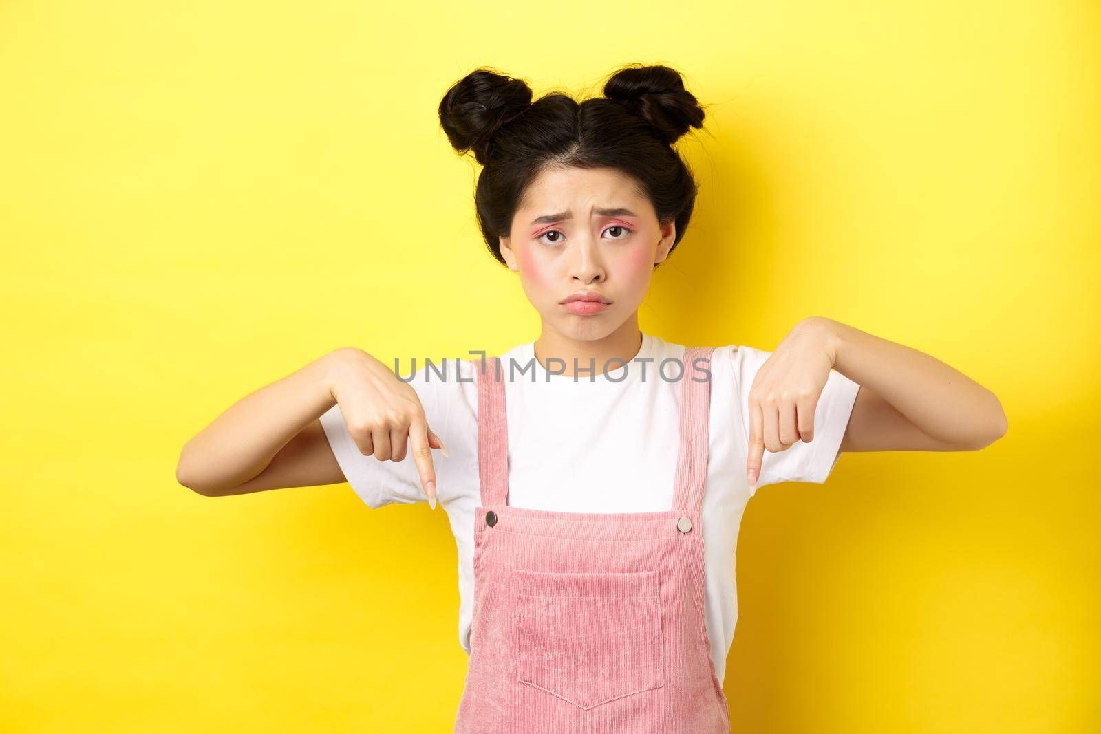 Sad frowning asian girl pointing fingers down at unfair thing, looking upset and gloomy, standing with makeup on yellow background.