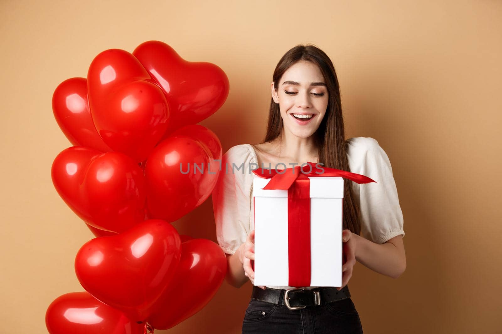 Happy woman open Valentines day gift, smiling excited and looking at present box, standing near red heart balloons on beige background by Benzoix