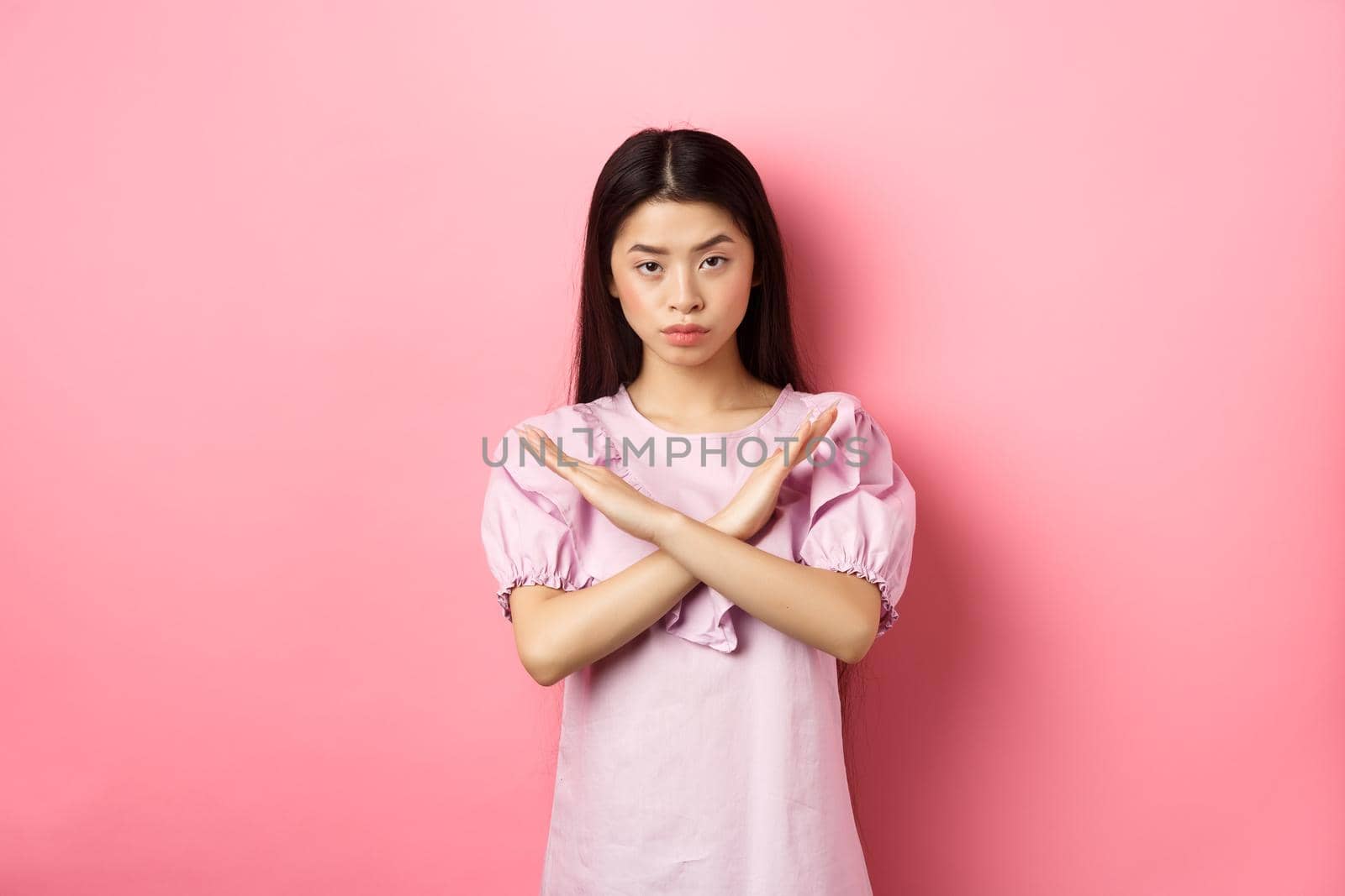 Serious chinese girl show cross sign, make stop gesture and look confident, block person, say no and reject offer, standing on pink background by Benzoix