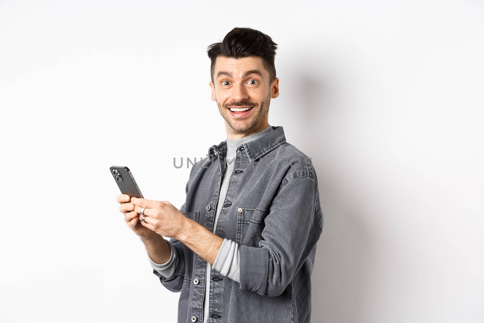 Handsome young man using smartphone and looking at camera. Caucasian guy chatting on phone with happy face, standing on white background.