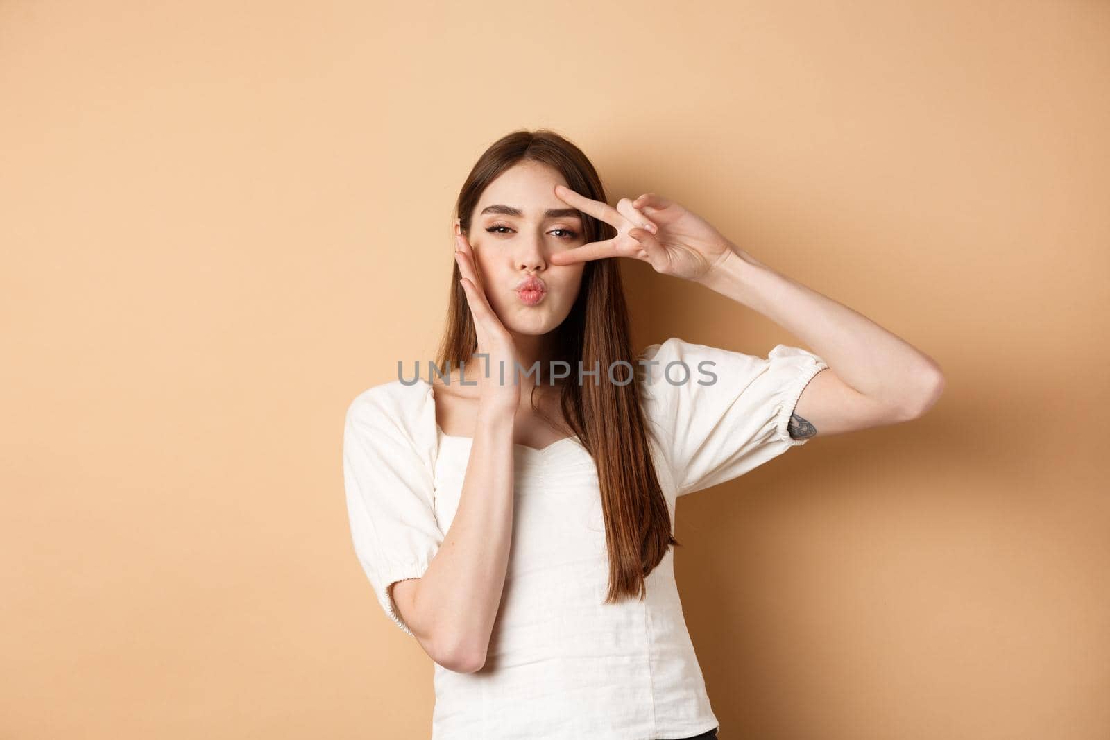 Cute woman showing v-sign and pucker lips for kiss, touching face with silly expression, standing on beige background.