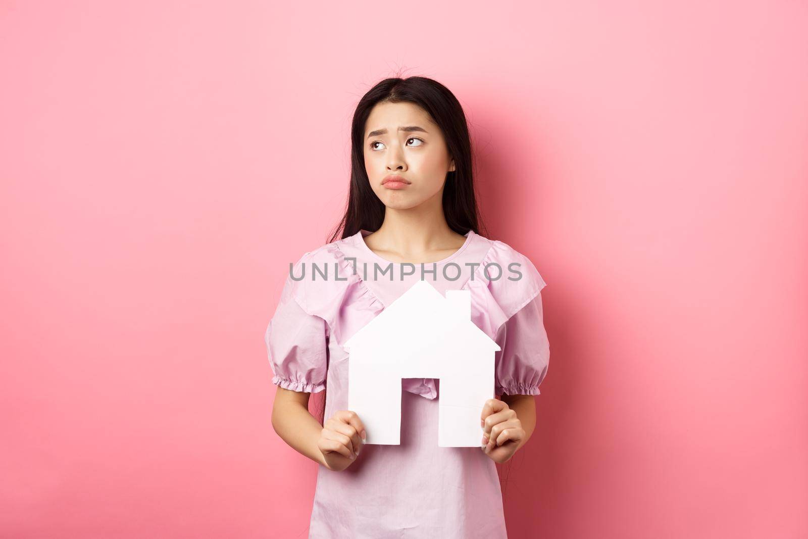 Real estate and insurance concept. Sad asian woman looking aside with unfair face, showing paper house cutout, dreaming of apartment, standing against pink background by Benzoix