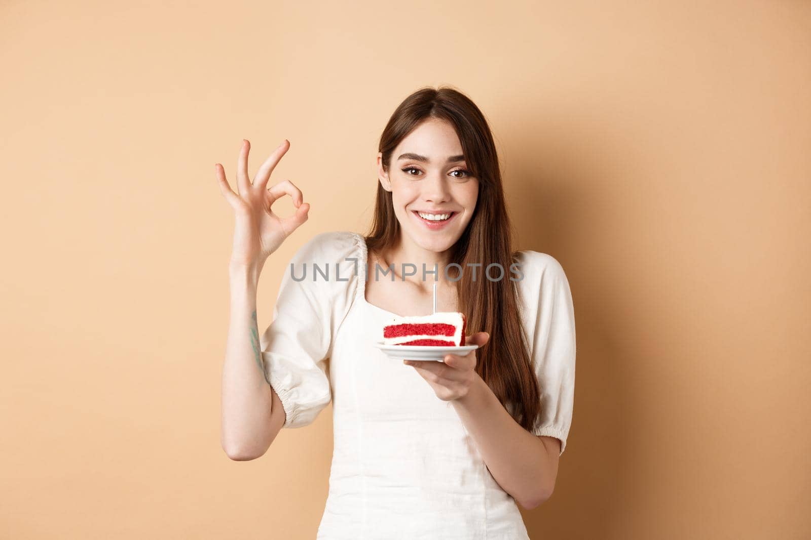 Happy birthday girl show okay gesture and hold bday cake, making wish on her holiday, standing on beige background.