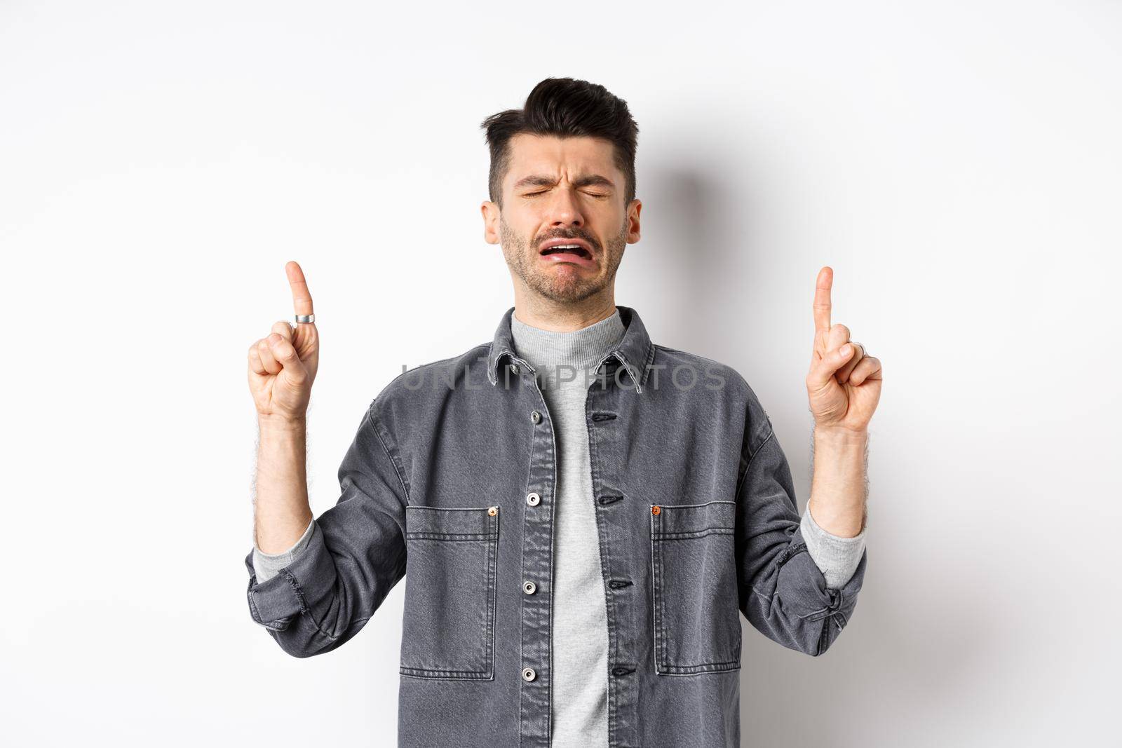 Sad and miserable guy crying while pointing fingers up, showing disappointed promo, sobbing and whining, standing on white background by Benzoix