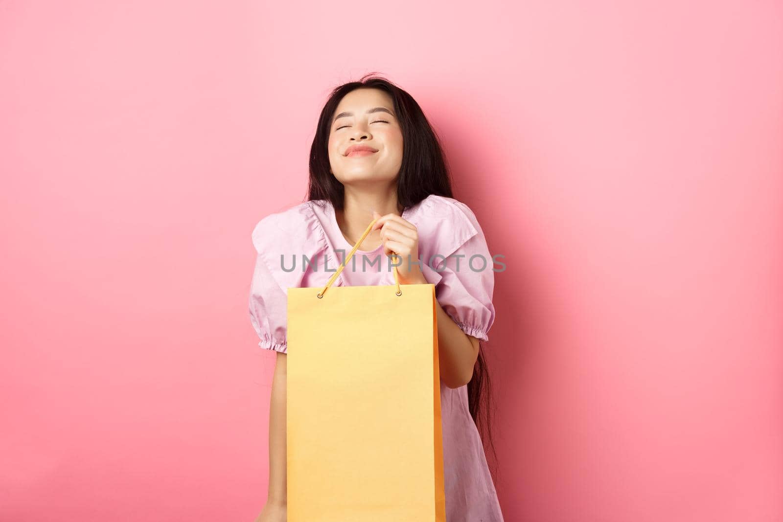 Beautiful happy asian woman holding shopping bag, smiling with eyes closed, standing in dress against pink background.
