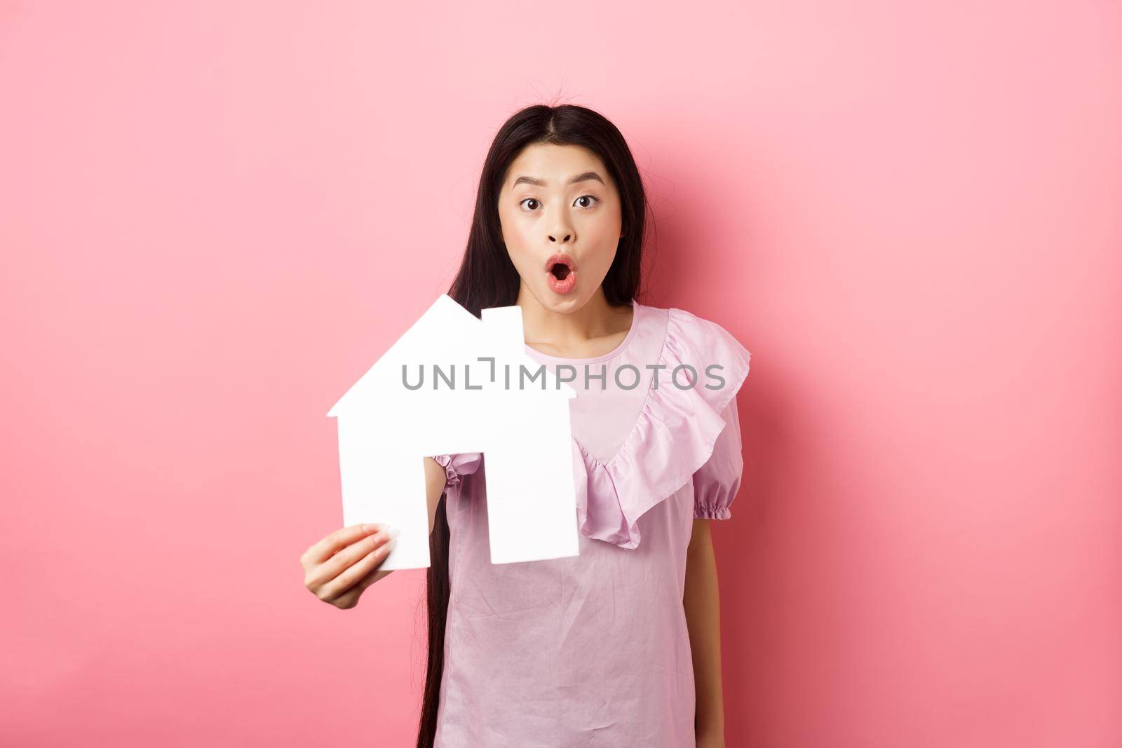 Real estate and insurance concept. Excited asian woman showing paper house cutout and look amazed, special offer, standing on pink background by Benzoix