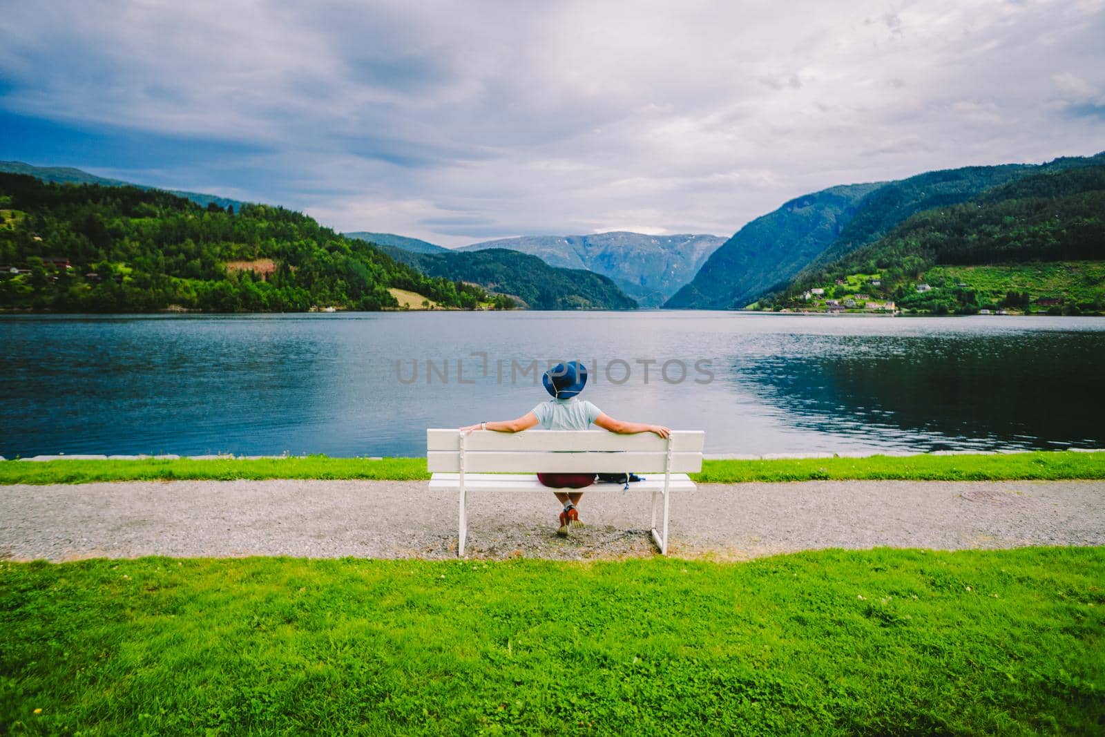 Woman sitting on a bench looking at the fjord in Ulvik, Norway. Fjord coastal promenade in Ulvik, Hordaland county, Norge. Lonely tourist in hat sits back on bench and admires scenery Scandinavia by Tomashevska
