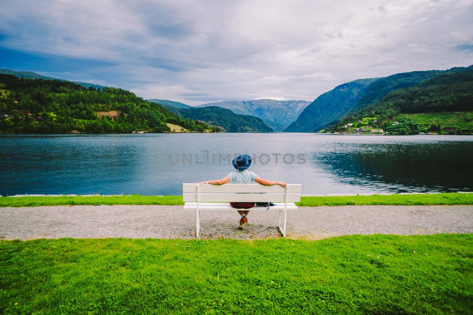 Woman sitting on a bench looking at the fjord in Ulvik, Norway. Fjord coastal promenade in Ulvik, Hordaland county, Norge. Lonely tourist in hat sits back on bench and admires scenery Scandinavia.