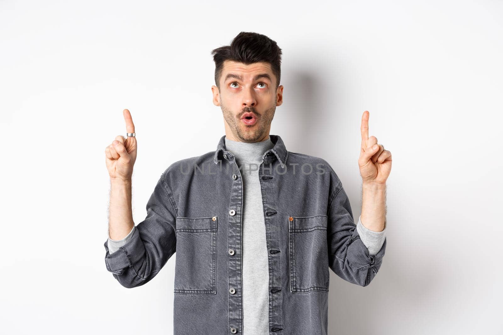 Excited modern man in denim jacket looking and pointing up, gasping amazed, checking out cool offer, standing on white background.