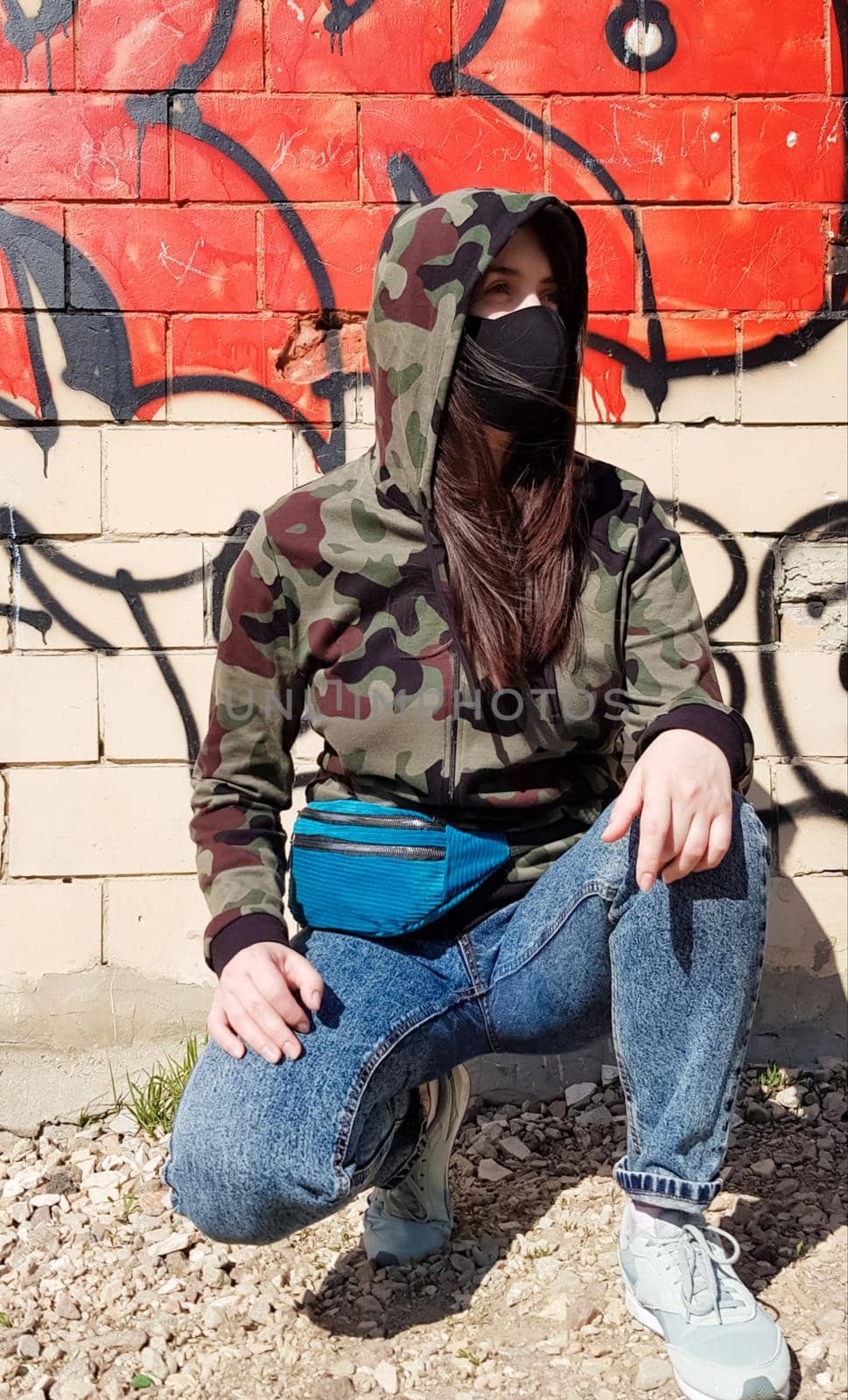 Ukraine, Kiev - April 24, 2020. A beautiful woman in a hood and a protective mask, dressed in blue jeans and a khaki camouflage jacket, sits near a wall with graffiti. On the street.