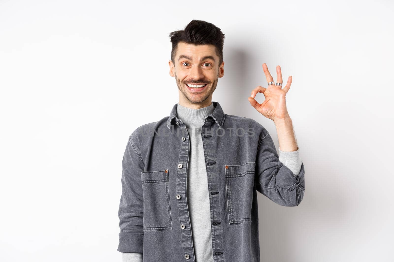 Smiling handsome man showing OK sign, like good product, recommending advertisement, standing satisfied on white background.