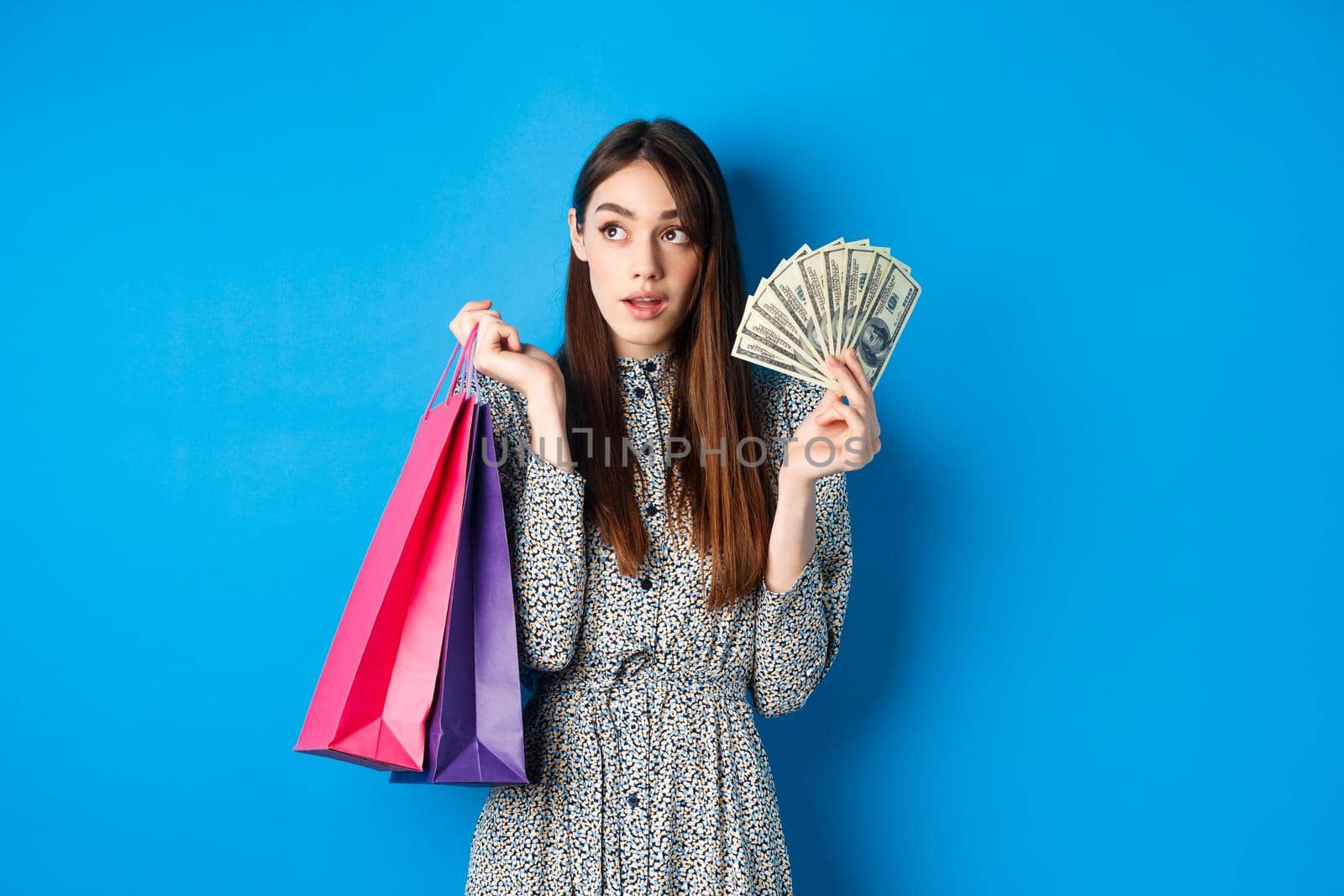 Shopping. Pretty woman thinking what to buy, looking left at logo, holding paper shop bags and money, standing on blue background.
