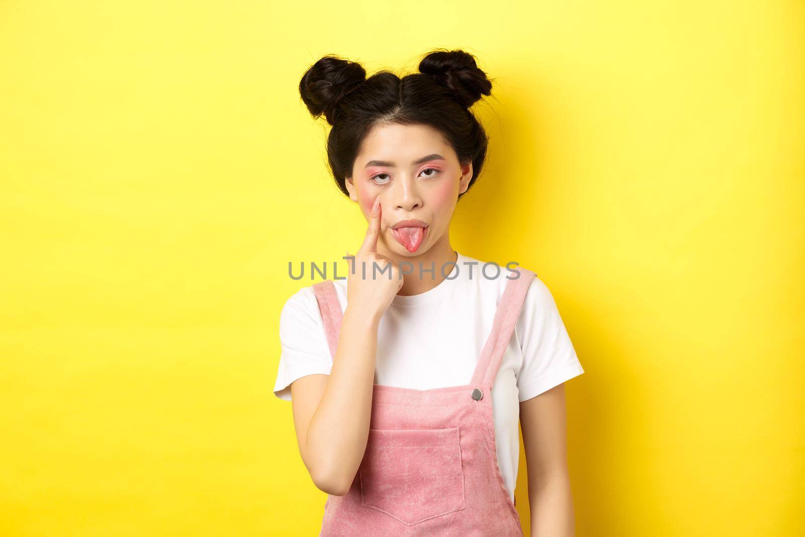 Rude asian girl stretch eyelid and showing tongue, mocking someone, standing on yellow background by Benzoix