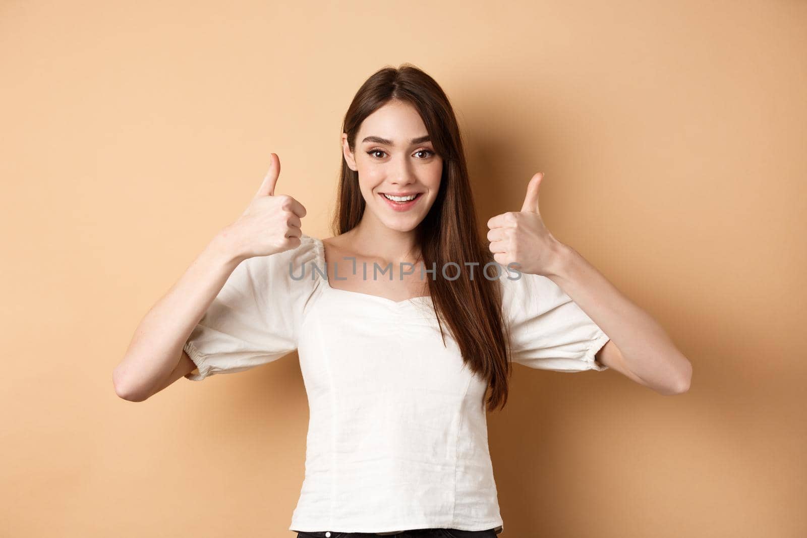 Very good. Happy and satisfied young woman showing thumbs-up, smiling positive, approve and like excellent choice, praising you, make well done gesture, beige background.
