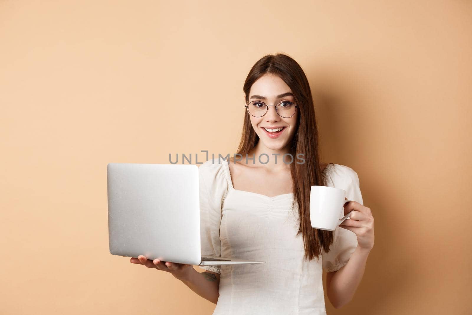 Cheerful woman in glasses drinking coffee and working on laptop, smiling at camera, standing on beige background.