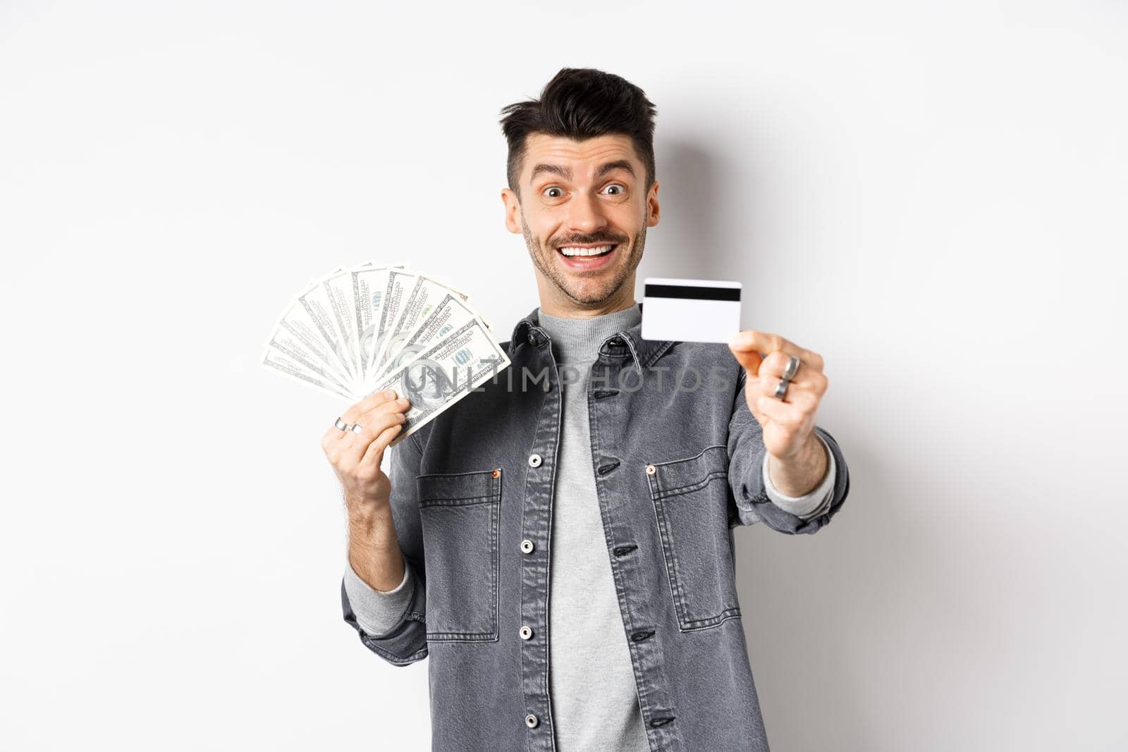 Image of handsome guy holding dollar bills but suggesting use plastic credit card, smiling friendly at camera, standing on white background.