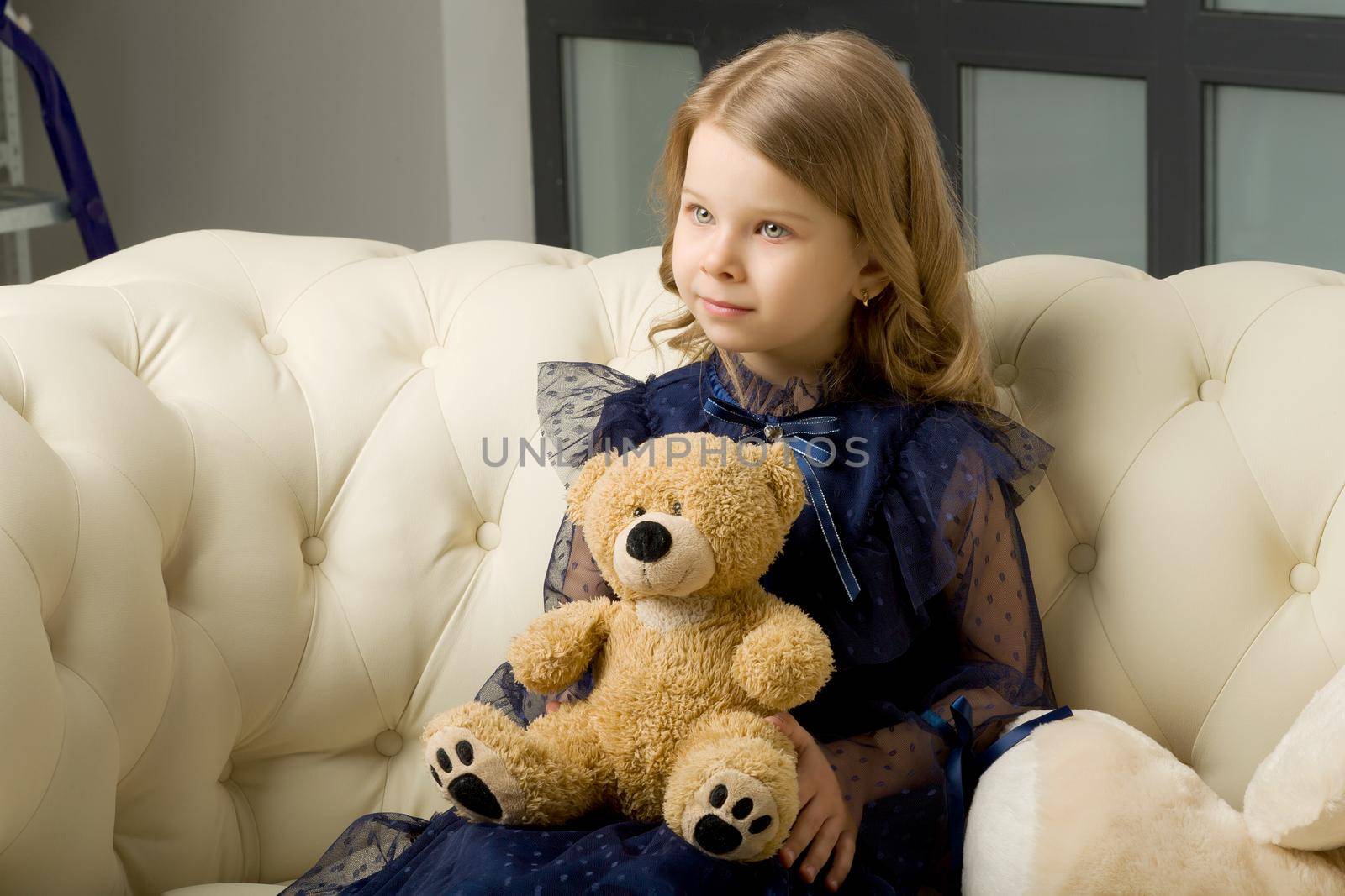 Portrait of girl with teddy bear sitting on sofa. Lovely blonde little girl wearing beautiful blue dress sitting on comfortable armchair in studio and looking away.